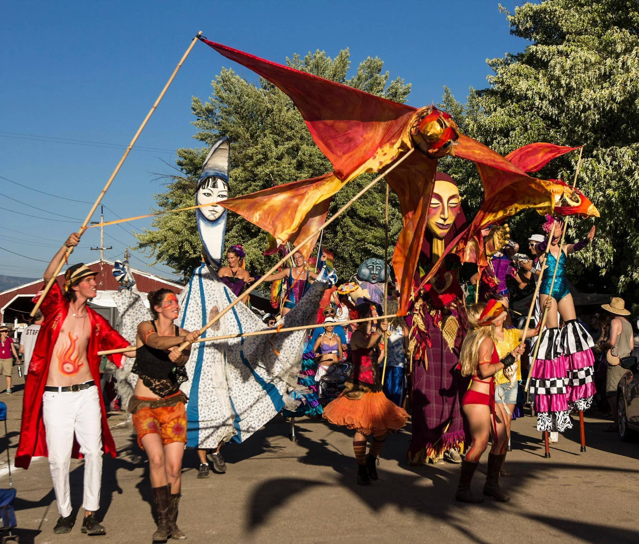 Street parade with performers in colorful costumes and large puppet-like figures holding orange and red fabric decorations