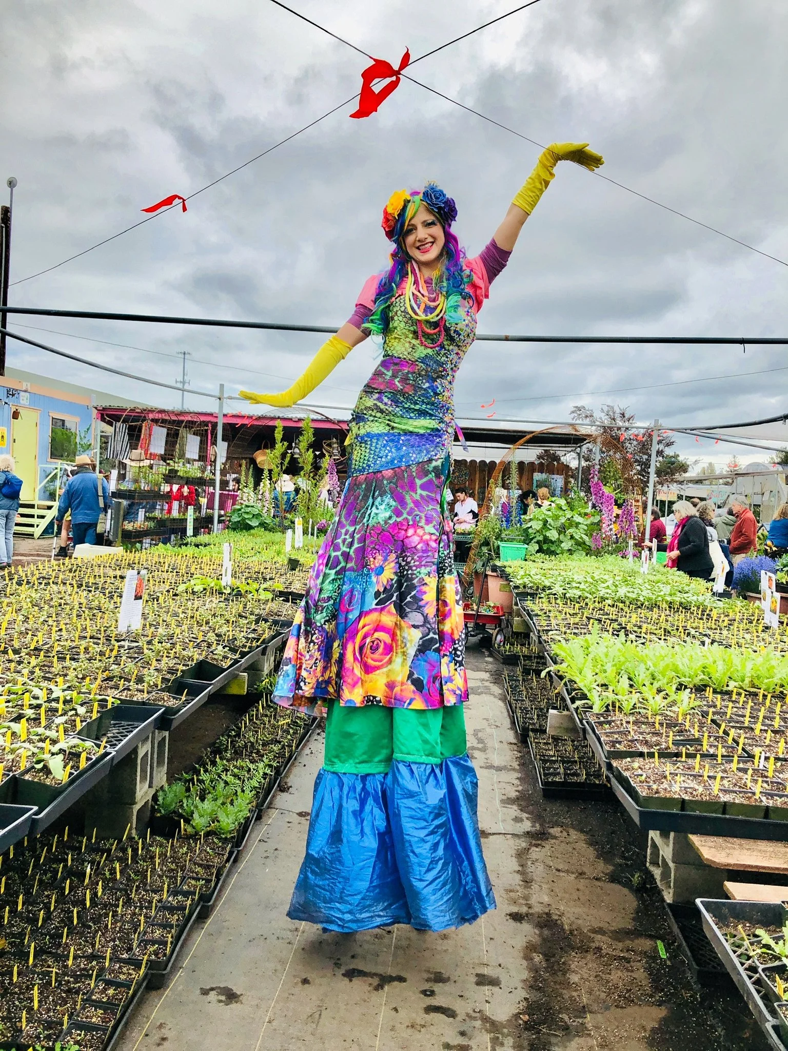 Woman dressed in colorful, eclectic outfit performing on stilts at a garden center with plants and customers in the background.