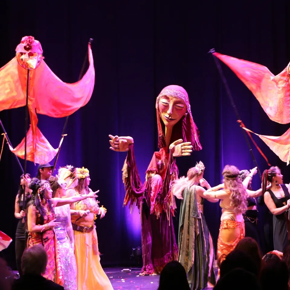 A theatrical stage performance featuring a large puppet with a feminine face and long purple hair, surrounded by performers holding large orange flags. The performers are dressed in colorful, whimsical costumes and are engaged in a dance or theatrica