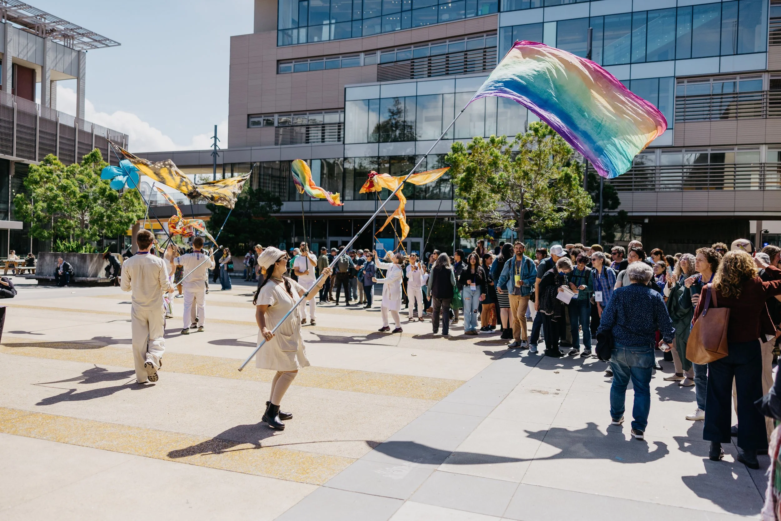 People gathered outdoors in an urban plaza with modern buildings. Two individuals are holding large, colorful, rainbow fabric banners.