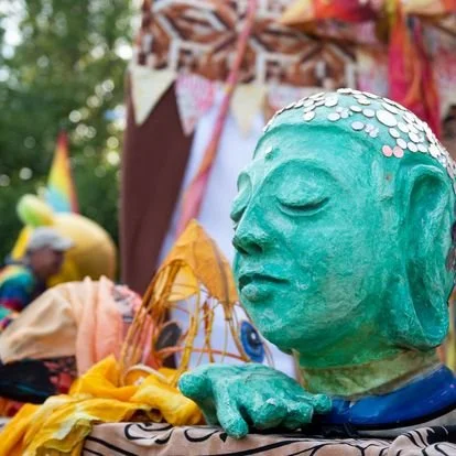 Close-up of a teal-colored sculpture of a person's head with closed eyes, adorned with white shell-like decorations on top. In the background, people and decorations are visible at what appears to be a festival or outdoor event.