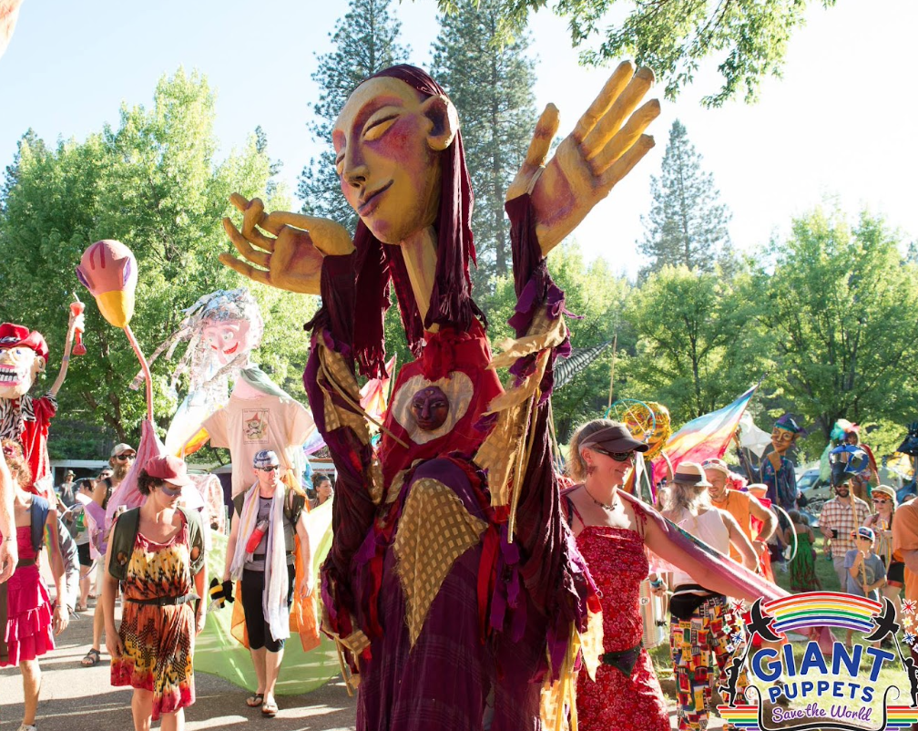 Colorful parade with giant puppet figures, including a central figure with a large smiling face, closed eyes, and outstretched arms, surrounded by people walking and dressed in vibrant outfits, in a park with green trees.
