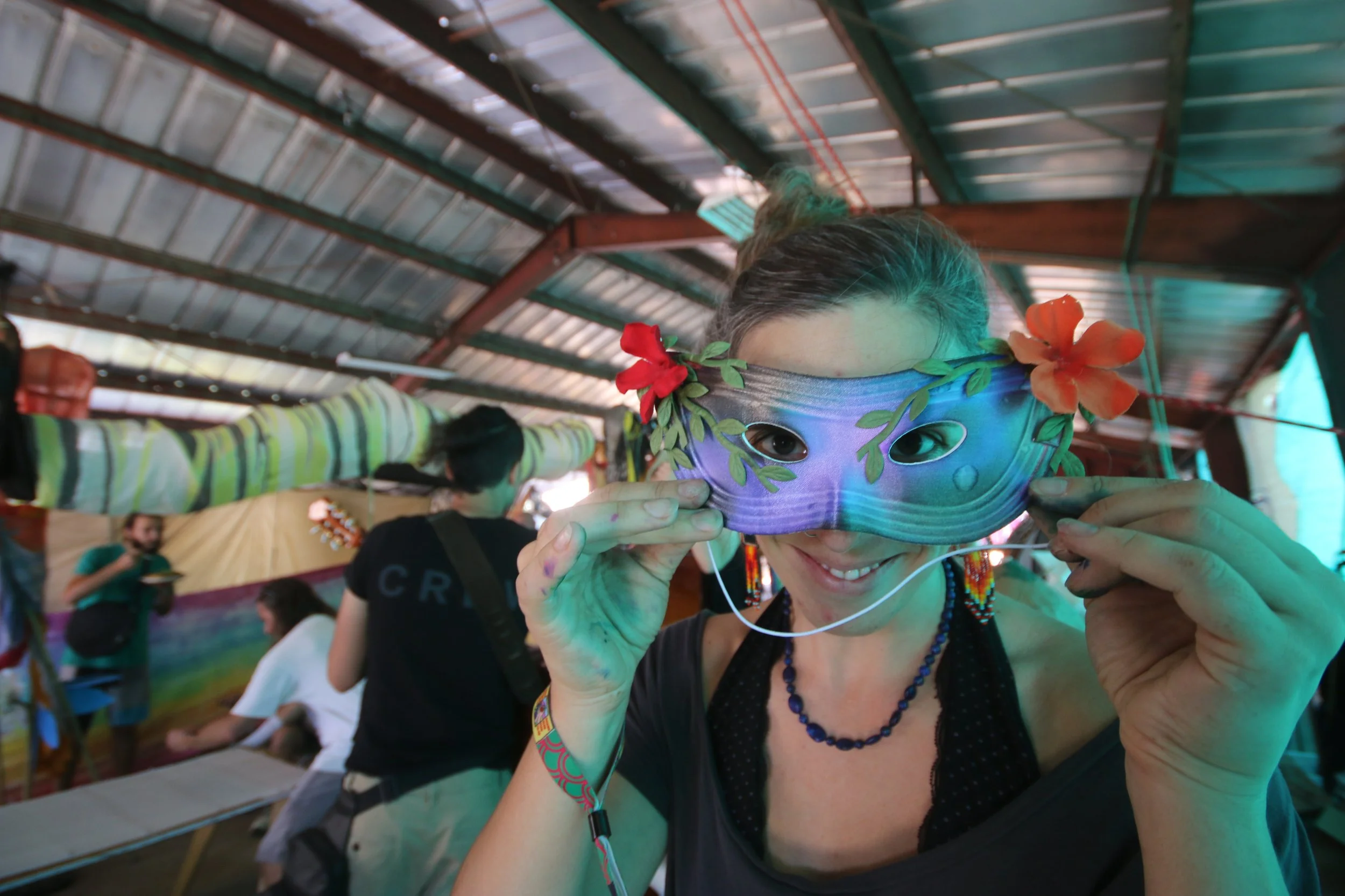 A woman smiling while holding a colorful mask with flowers over her eyes, at an indoor event with several people in the background.