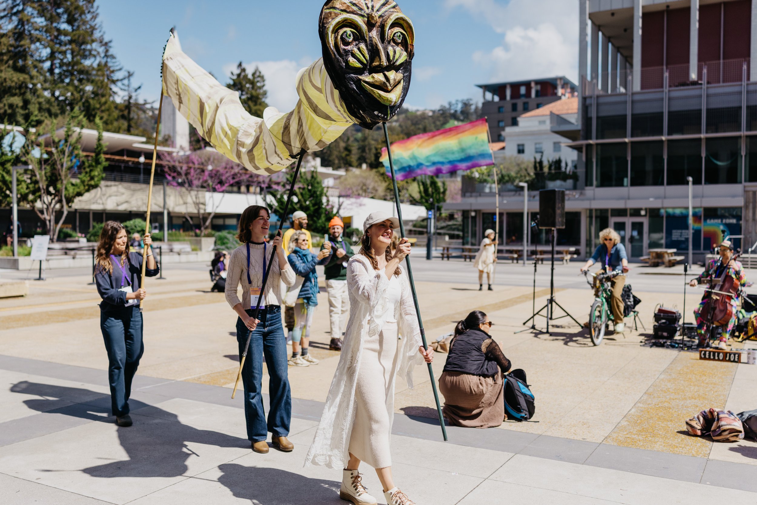 People participating in a pride parade, holding rainbow flags and puppet balloons, with colorful rainbow banners, in an outdoor urban plaza setting.