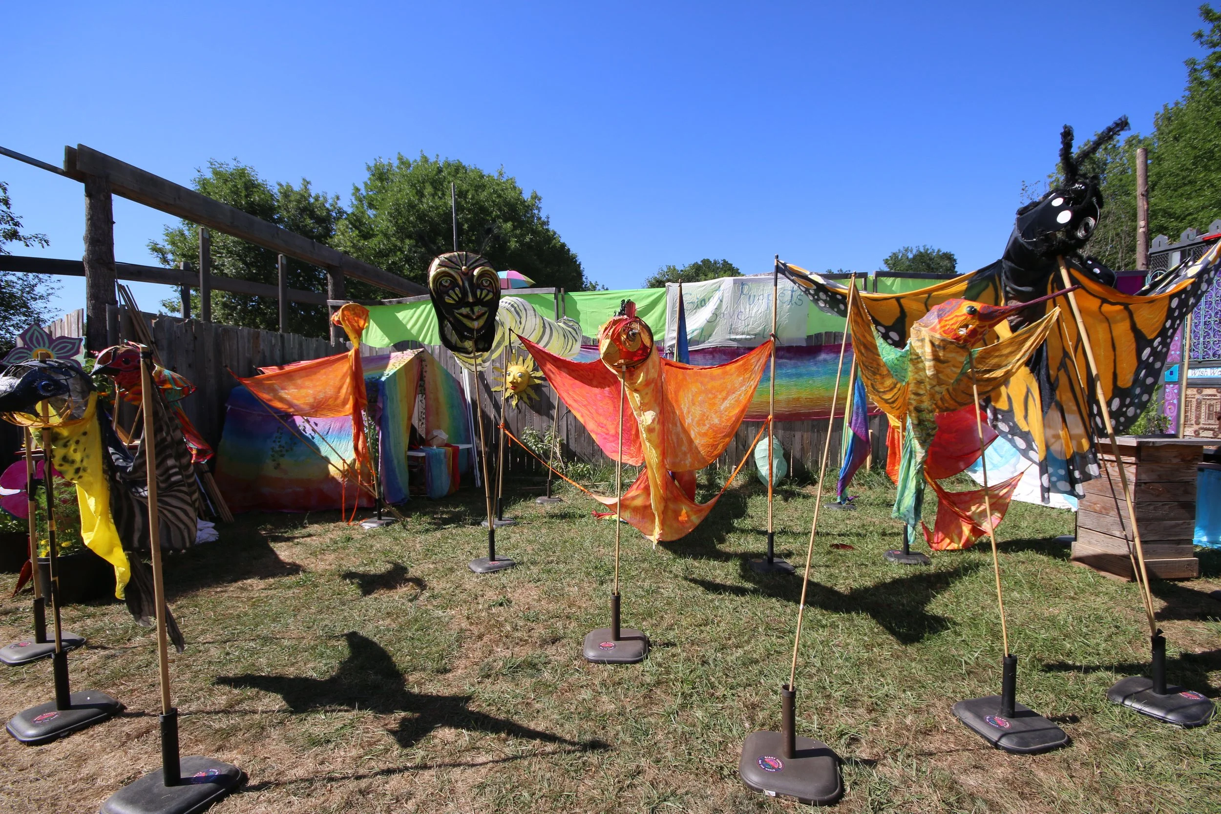 Colorful butterfly and insect-themed kites displayed outdoors on a grassy area with a wooden fence and trees in the background.