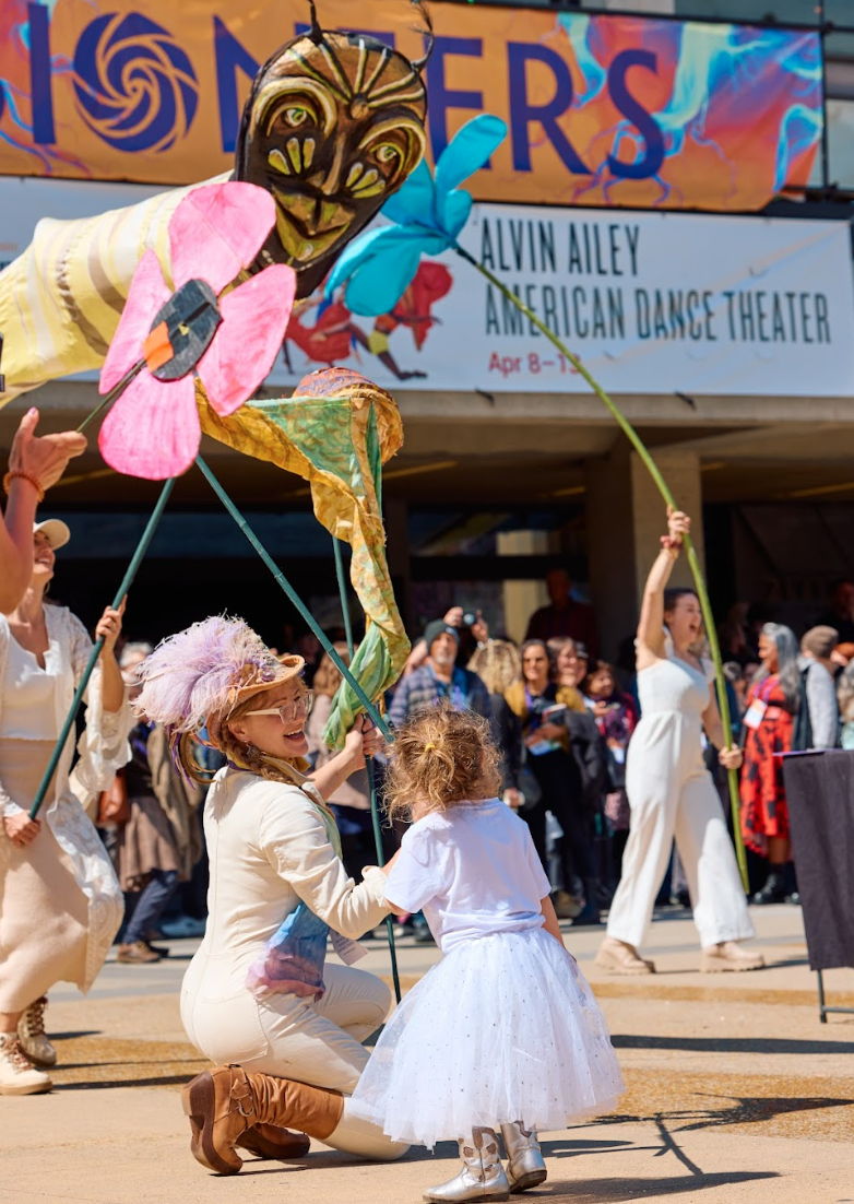 People participating in a parade or festival, holding colorful butterfly and flower puppets, with spectators watching in the background, and a banner advertising a dance theater event.