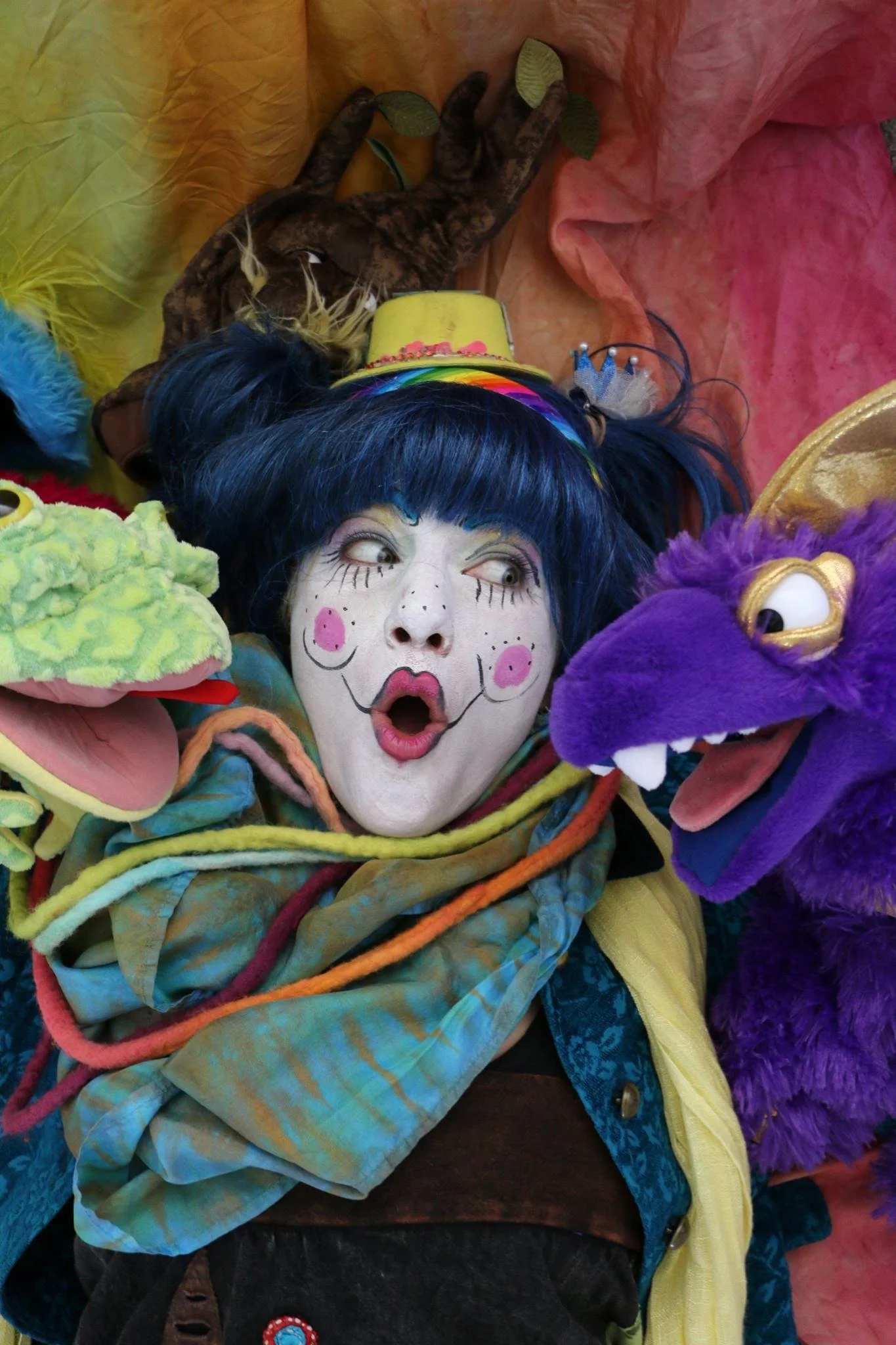 A person dressed as a clown with exaggerated makeup, blue hair, and colorful clothing is surrounded by plush puppets of a green frog and a purple dragon, with a background of bright, crinkled fabric.