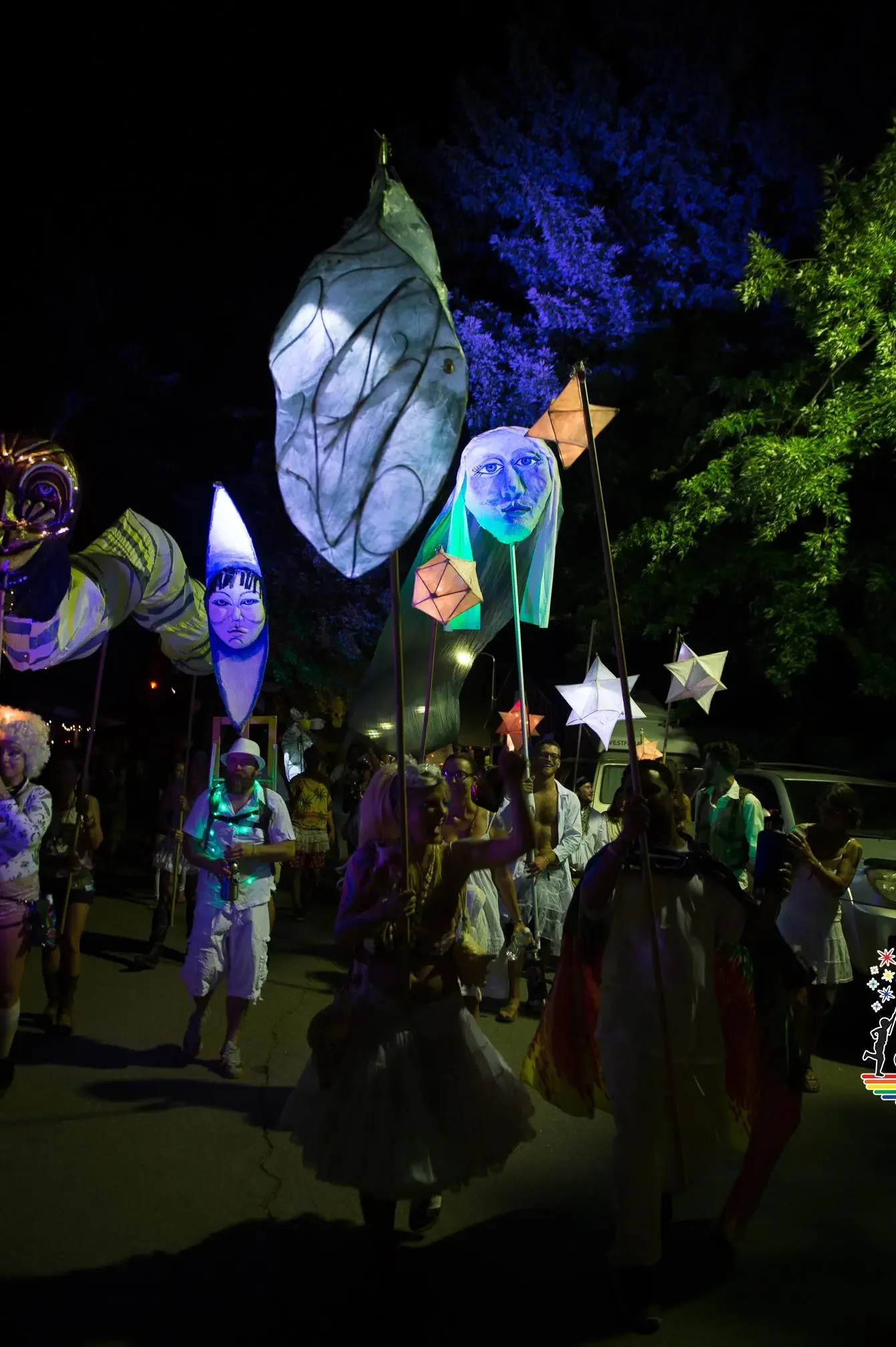 Nighttime parade with illuminated lanterns shaped like faces and stars, people dressed in costumes, and a large glowing leaf-shaped lantern, with trees in the background.