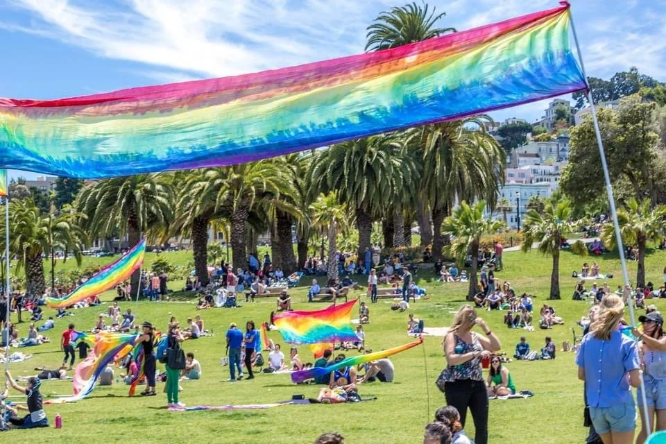 People flying rainbow-colored kites in a park with palm trees and buildings in the background on a sunny day.