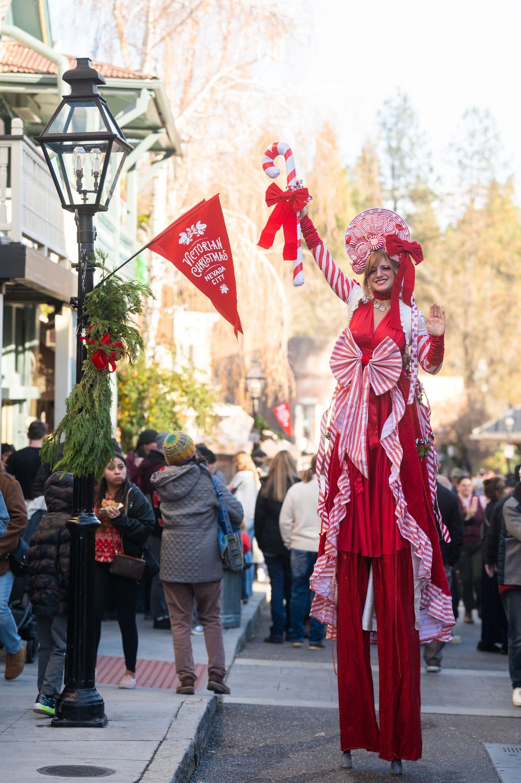 A woman on stilts dressed in red and white Christmas-themed costume with big bow, candy cane prop, and lollipop headpiece at a Christmas event in a city street, holding a flag that reads "Victorian Christmas Nevada City."