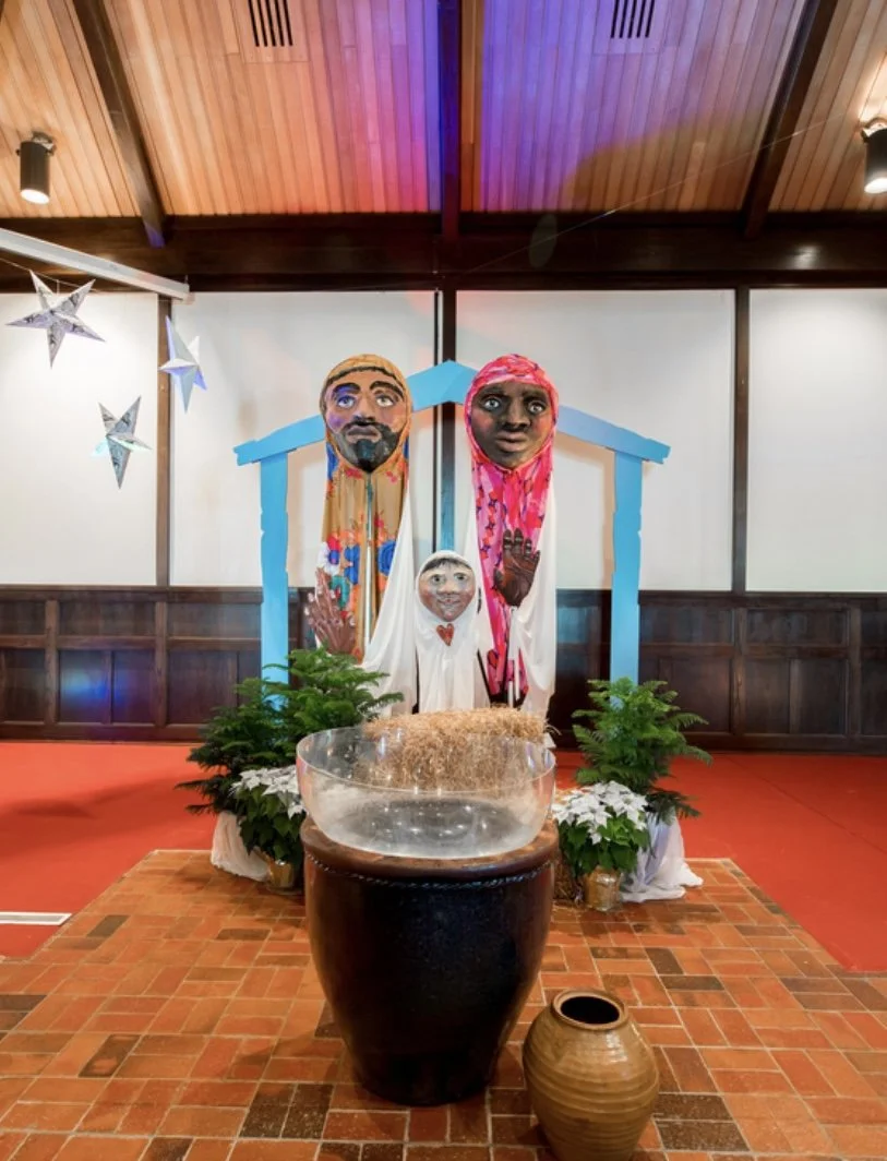 Colorful Nativity scene with three large papier-mâché faces representing Mary, Joseph, and baby Jesus, surrounded by green plants, a water basin, and a clay pot, inside a wooden-paneled room.