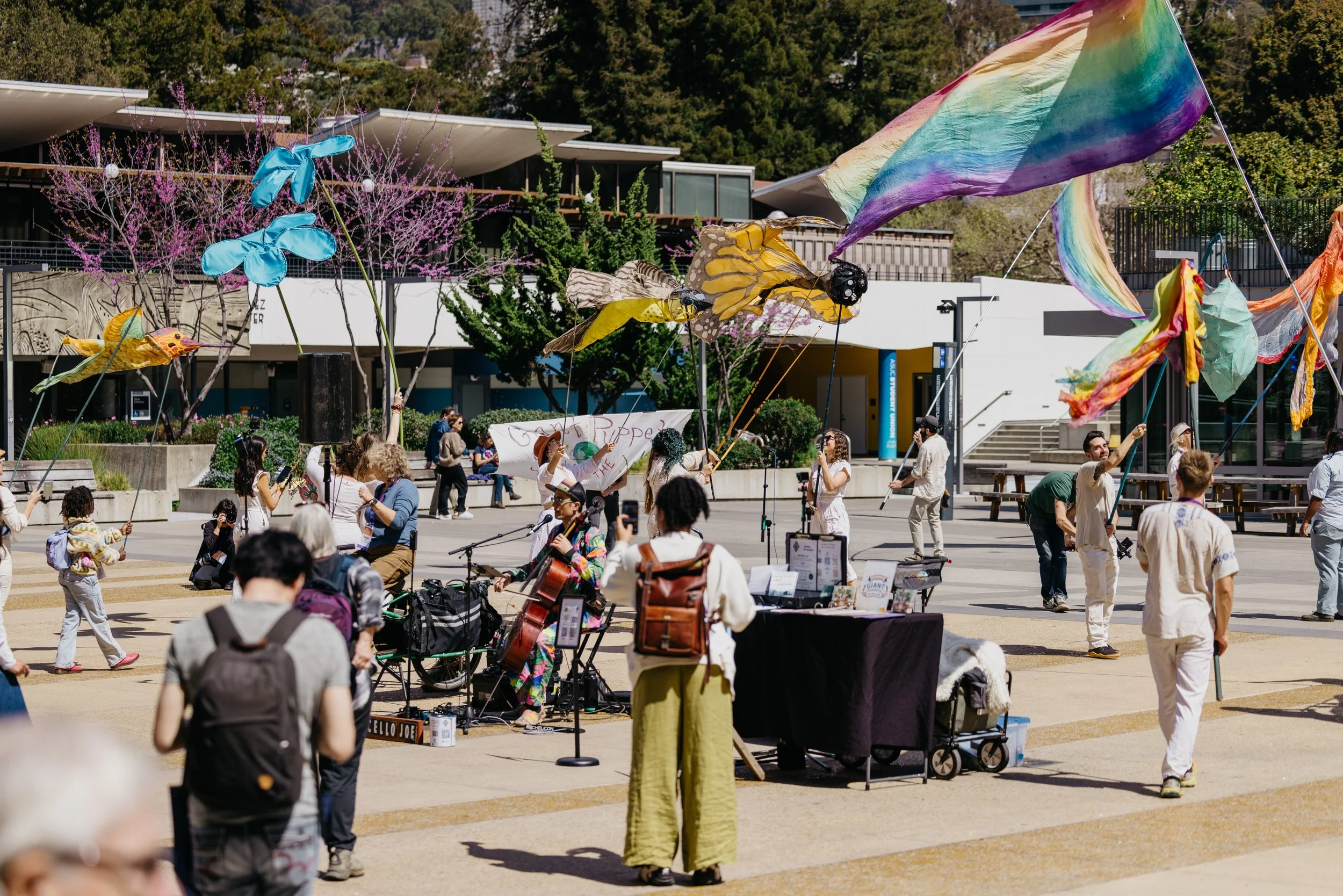 People gathering outdoors at a community event with colorful flags, banners, and a musician playing guitar with a microphone, surrounded by trees in bloom and modern buildings.