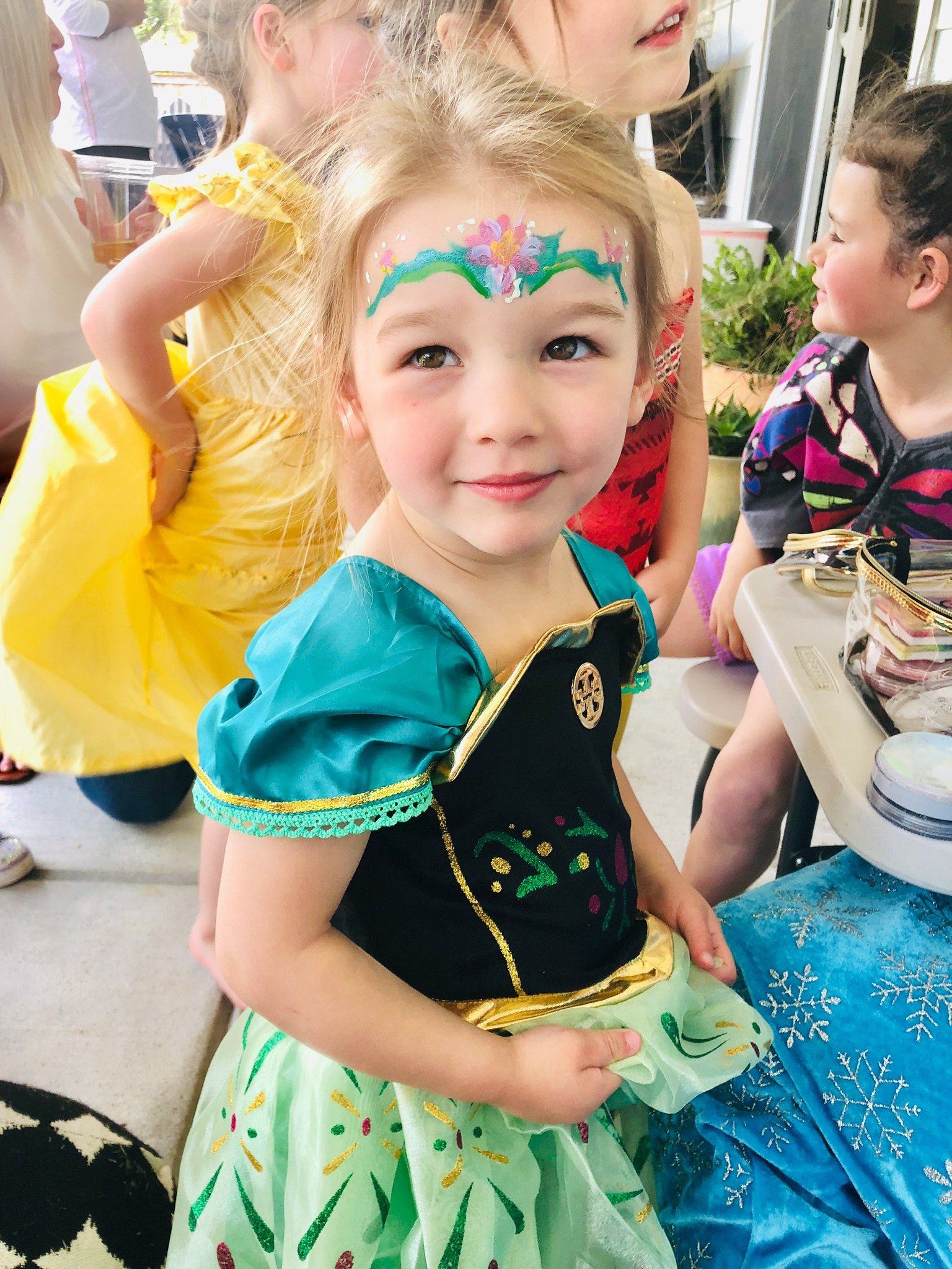 A young girl dressed in a princess costume with face paint of a colorful flower crown, standing with other children in a festive setting.