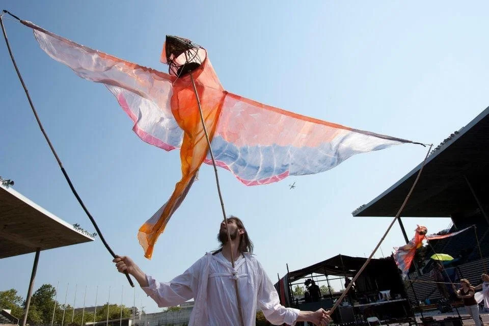 Person flying a large colorful kite with wings resembling a bird, in an outdoor area during the daytime.