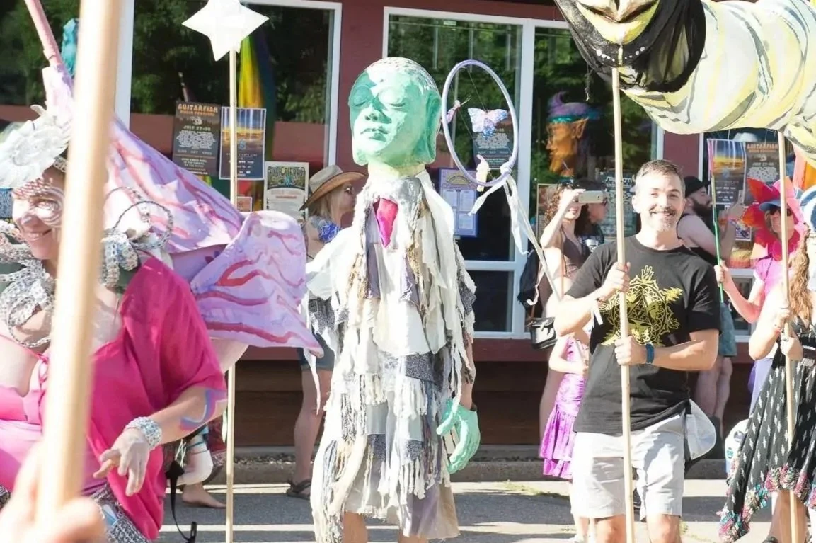 People participating in a colorful parade, dressed in costumes with large hats, masks, and artistic outfits, some holding balloons and props, outside a building.
