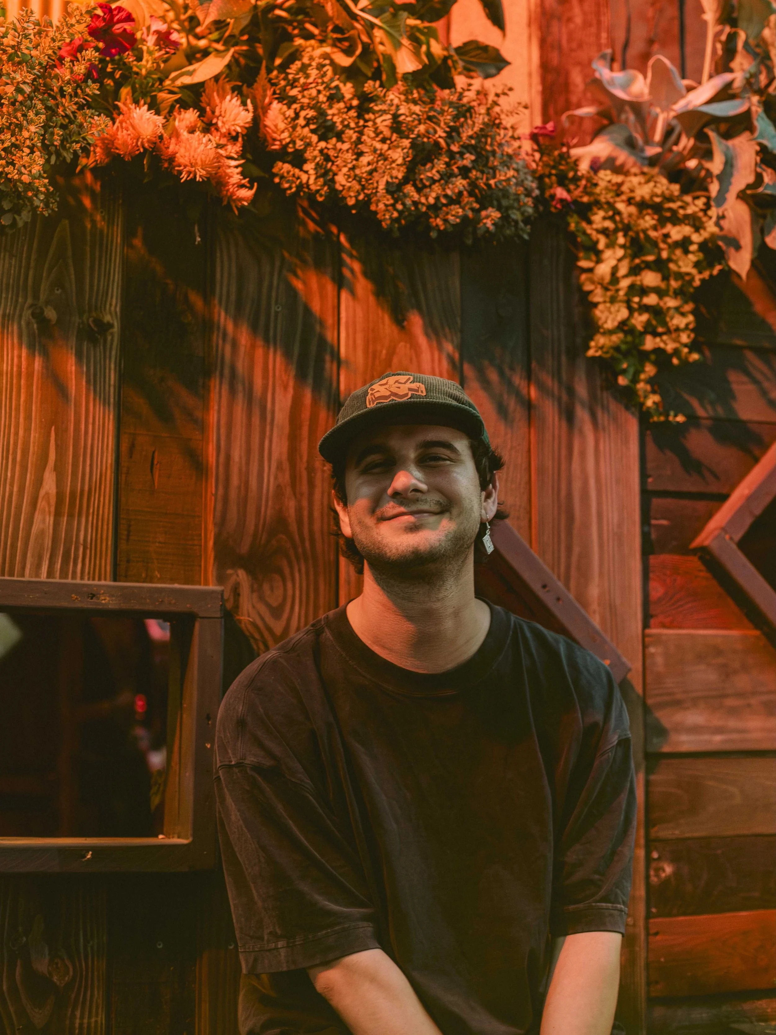 A smiling young man in a black t-shirt and baseball cap sitting in front of a wooden wall decorated with flowers and greenery.