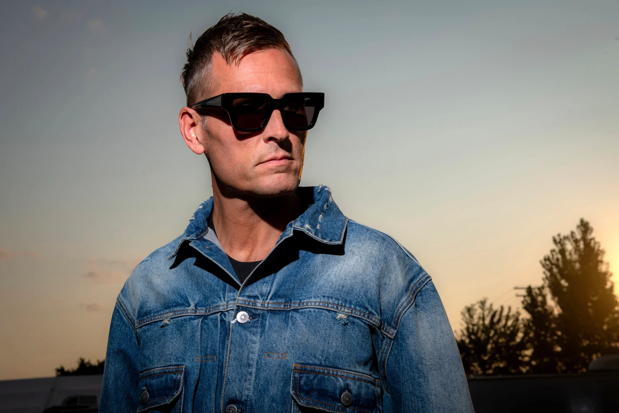 Man wearing black sunglasses and a denim jacket standing outdoors during sunset or sunrise with trees in the background.