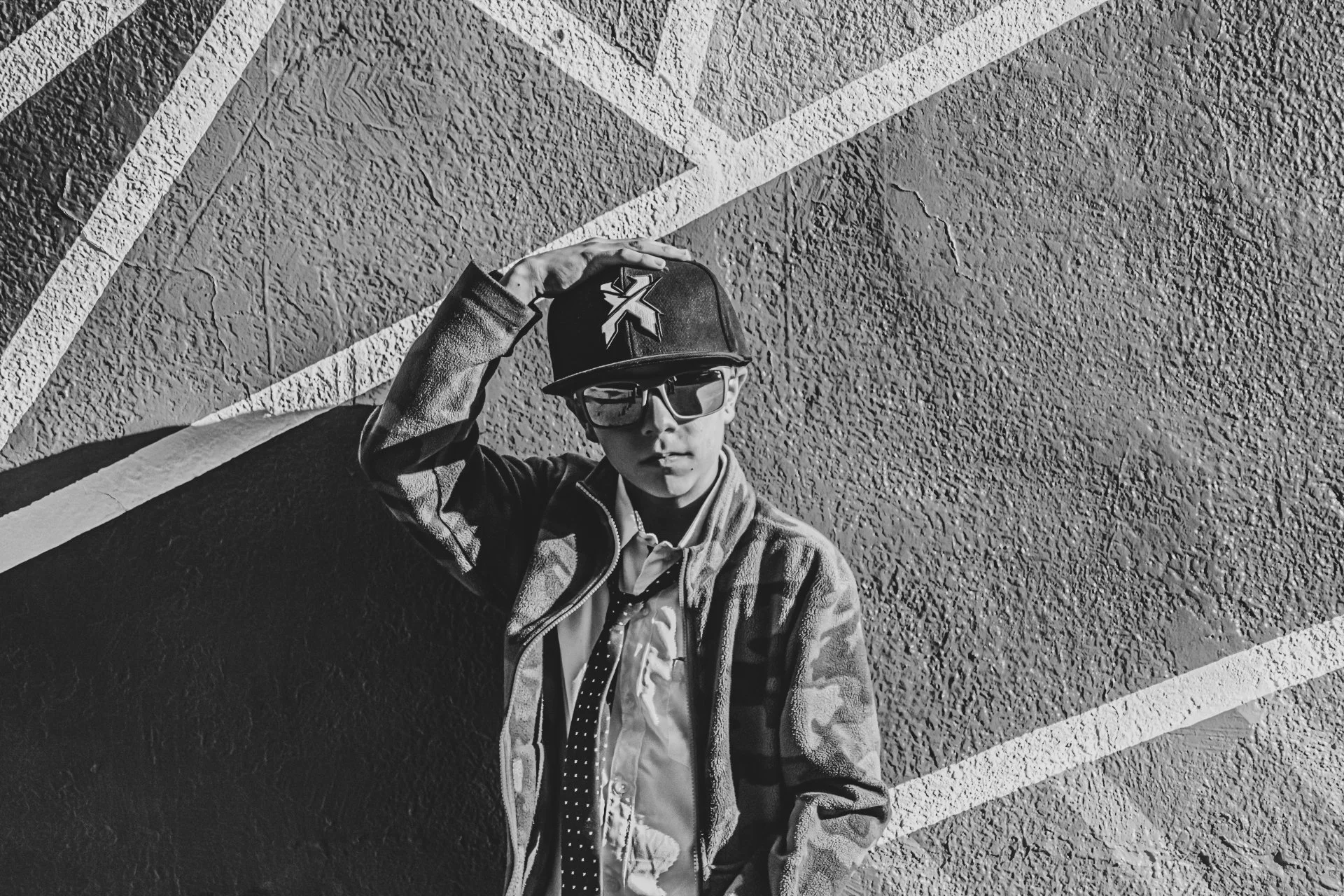 A young man wearing a cap with a star logo, sunglasses, a camouflage jacket, and a collared shirt with a dotted tie. He is standing near a textured wall with diagonal lines and is touching his cap with one hand.