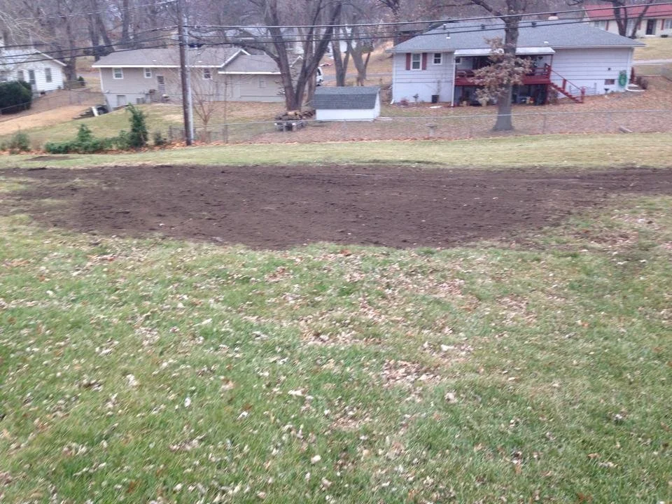 Backyard with a freshly tilled patch of soil and surrounding grass, suburban homes and leafless trees in the background.