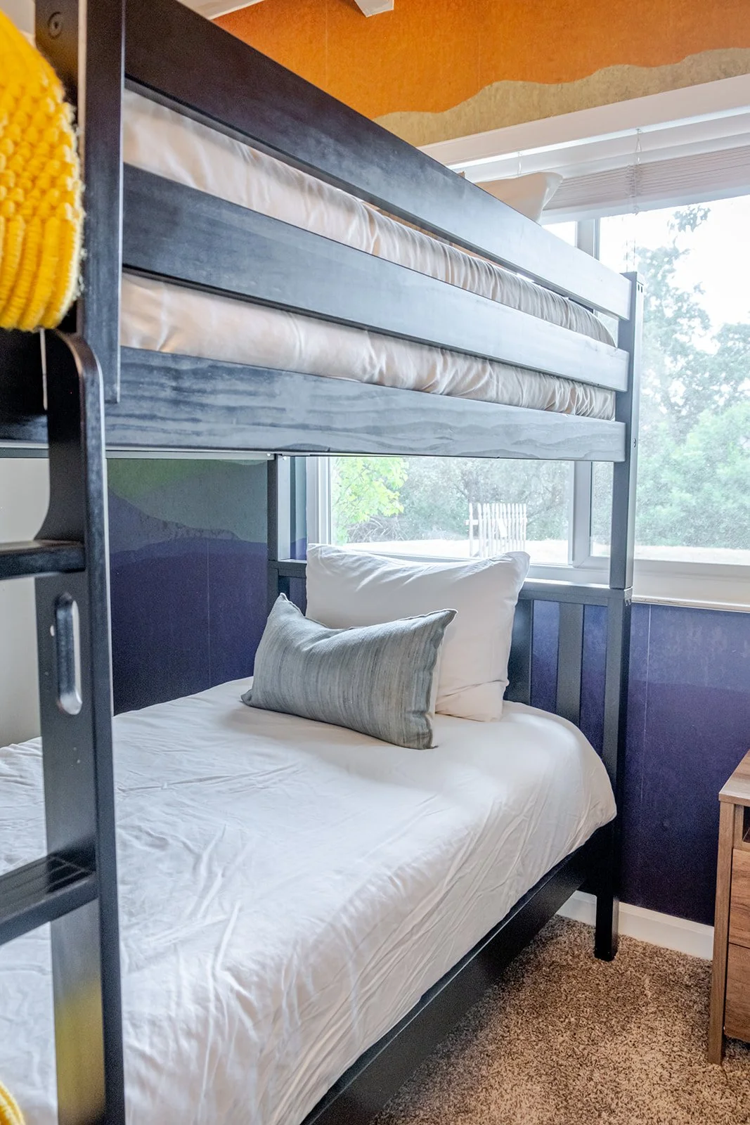 Bunk beds with a window view of trees, the top bunk has a mattress and bed linens, and the bottom bunk has a pillow and white sheet.