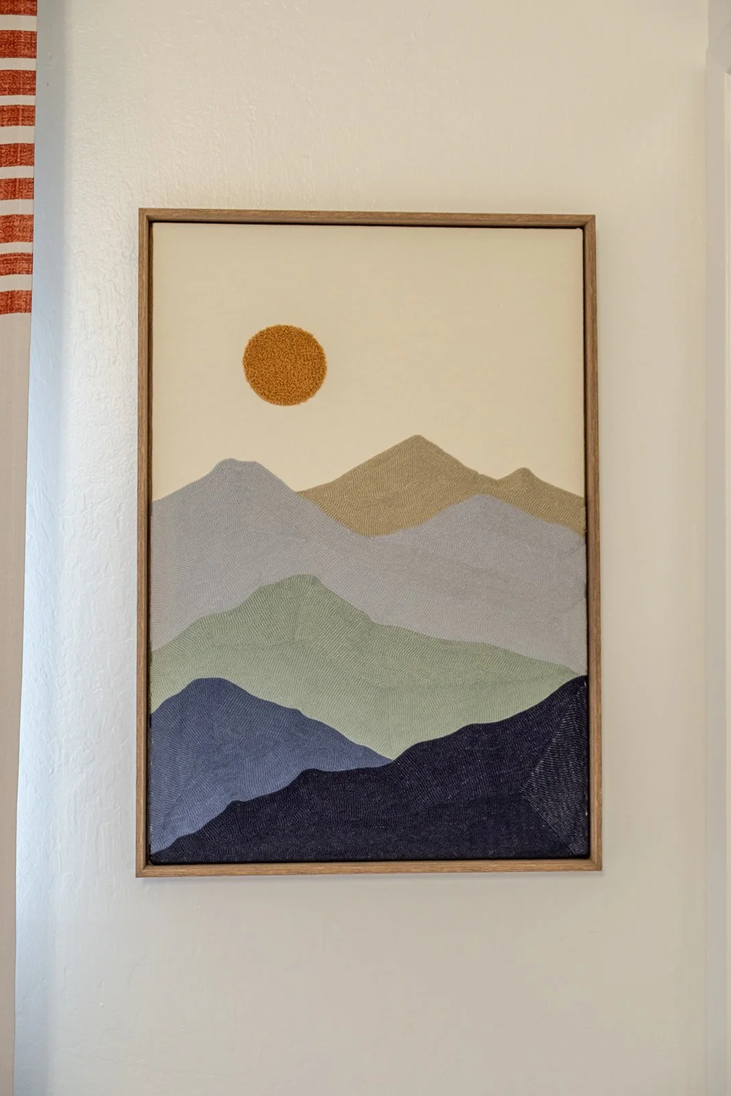A framed textile art piece depicting a mountain landscape with layered hills in shades of gray, green, and blue, and an orange sun against a beige background.