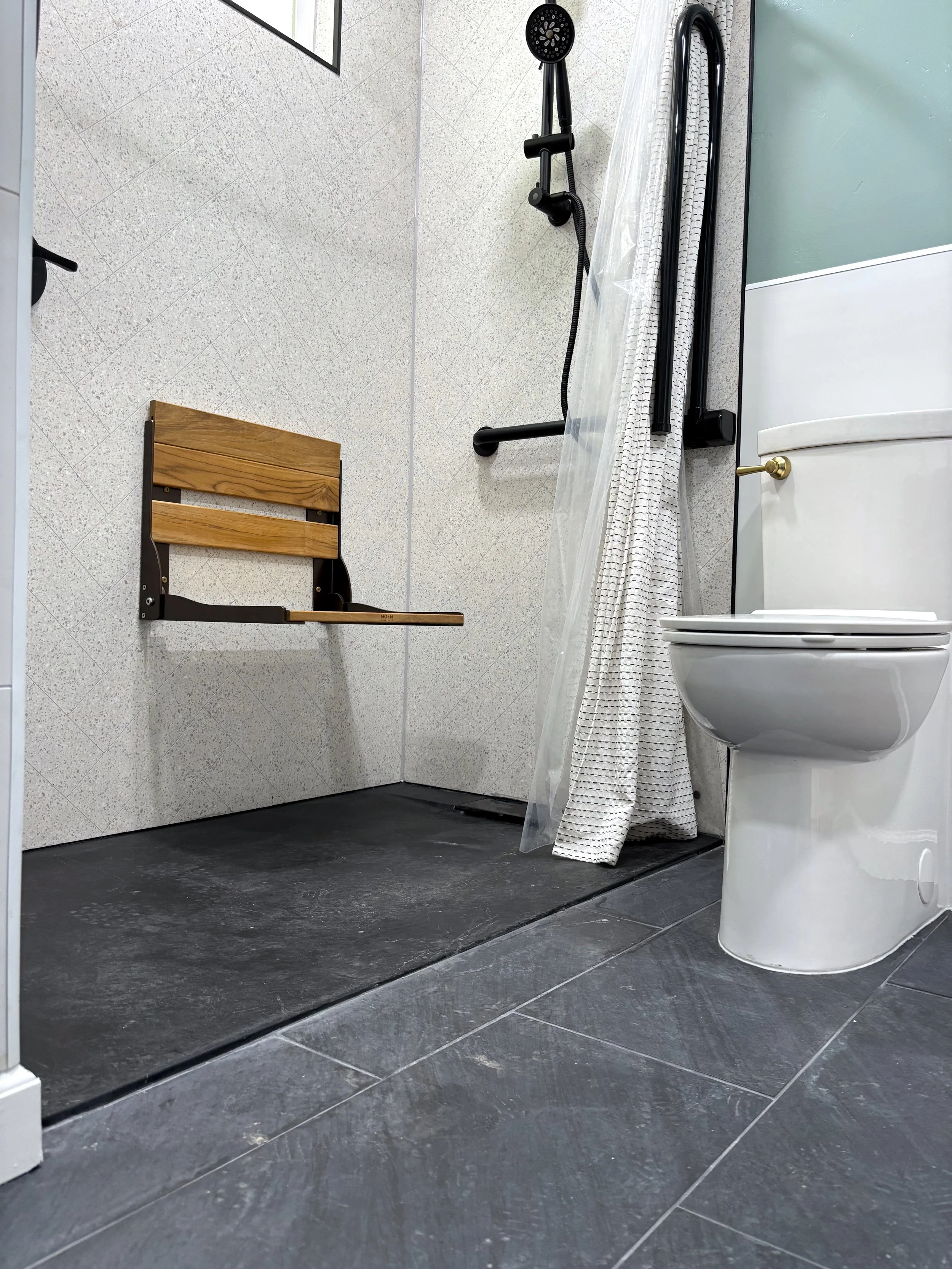 An empty walk-in shower with a wooden fold-down seat, black grab bars, a handheld showerhead, and a curtain. To the right, there is a white toilet with a gold handle. The bathroom has speckled, beige walls and dark gray tiles on the floor.