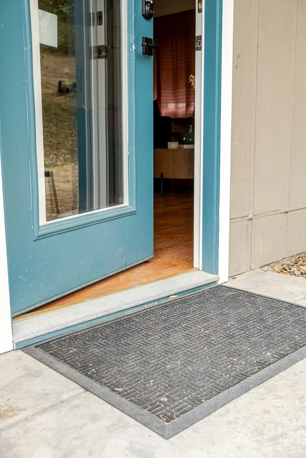 Open blue front door with glass panel, revealing a wooden floor and room with a cabinet and objects inside. Black rubber doormat with textured pattern outside the door.