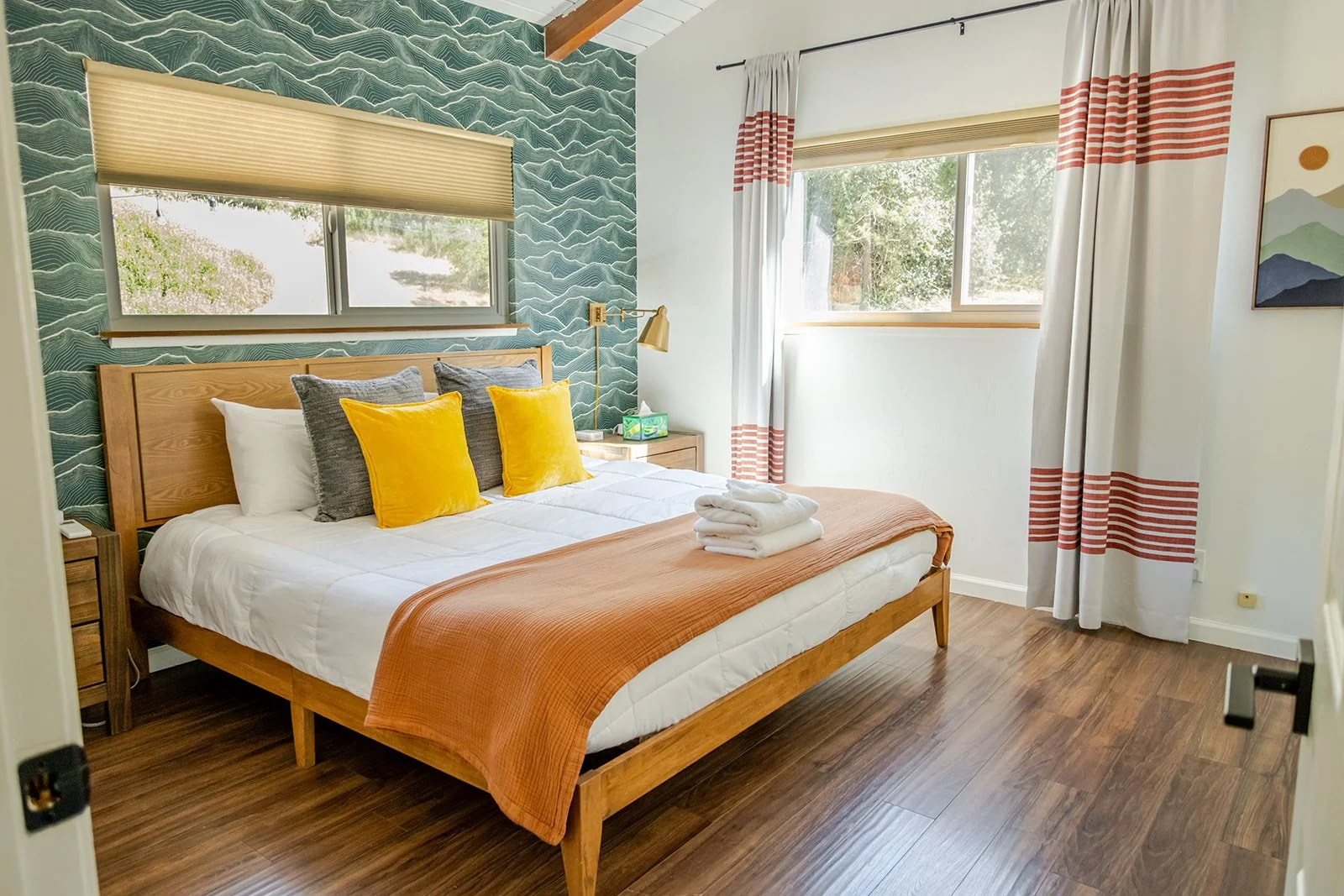 A bedroom with a wooden bed frame, white bedding, an orange bedspread, and four decorative pillows (two yellow and two gray). There are two windows, one with a beige blind and the other with white curtains with red stripes. The room has wooden floori