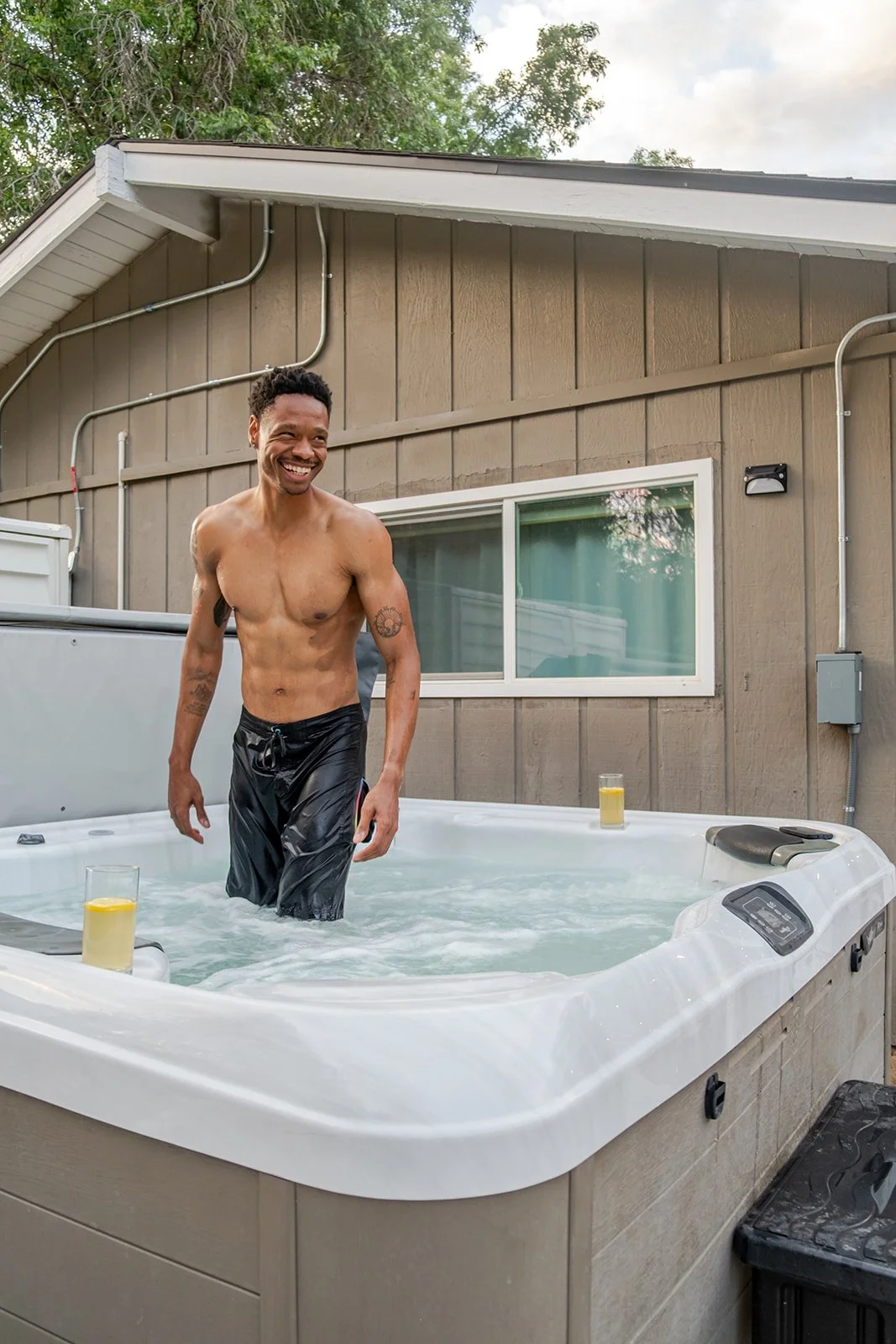 A smiling man with tattoos is standing in an outdoor hot tub, wearing black swim trunks. The hot tub has two glasses with lemon slices on its edge, and is situated on a deck next to a gray building with a window. There are trees and a cloudy sky in t
