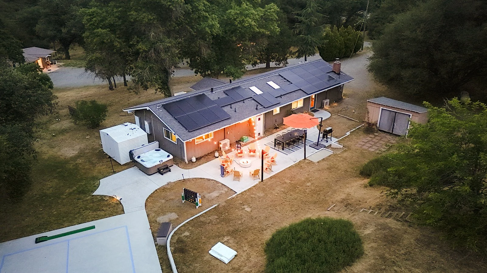 An aerial view of a house with solar panels on the roof during dusk, featuring a backyard patio with outdoor furniture, an umbrella, a fire pit with chairs, and a hot tub, surrounded by trees and neighboring houses.