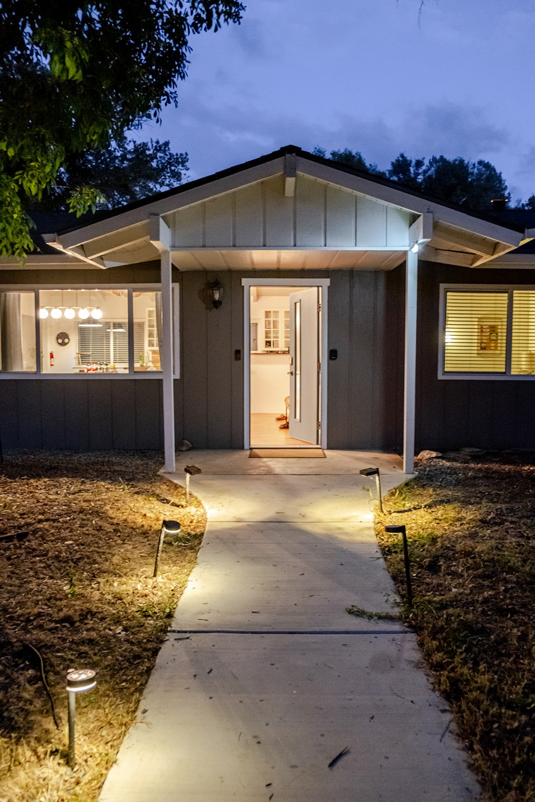 Front view of a house at dusk with illuminated walkway leading to the open front door, visible interior through window, trees and cloudy sky in the background.
