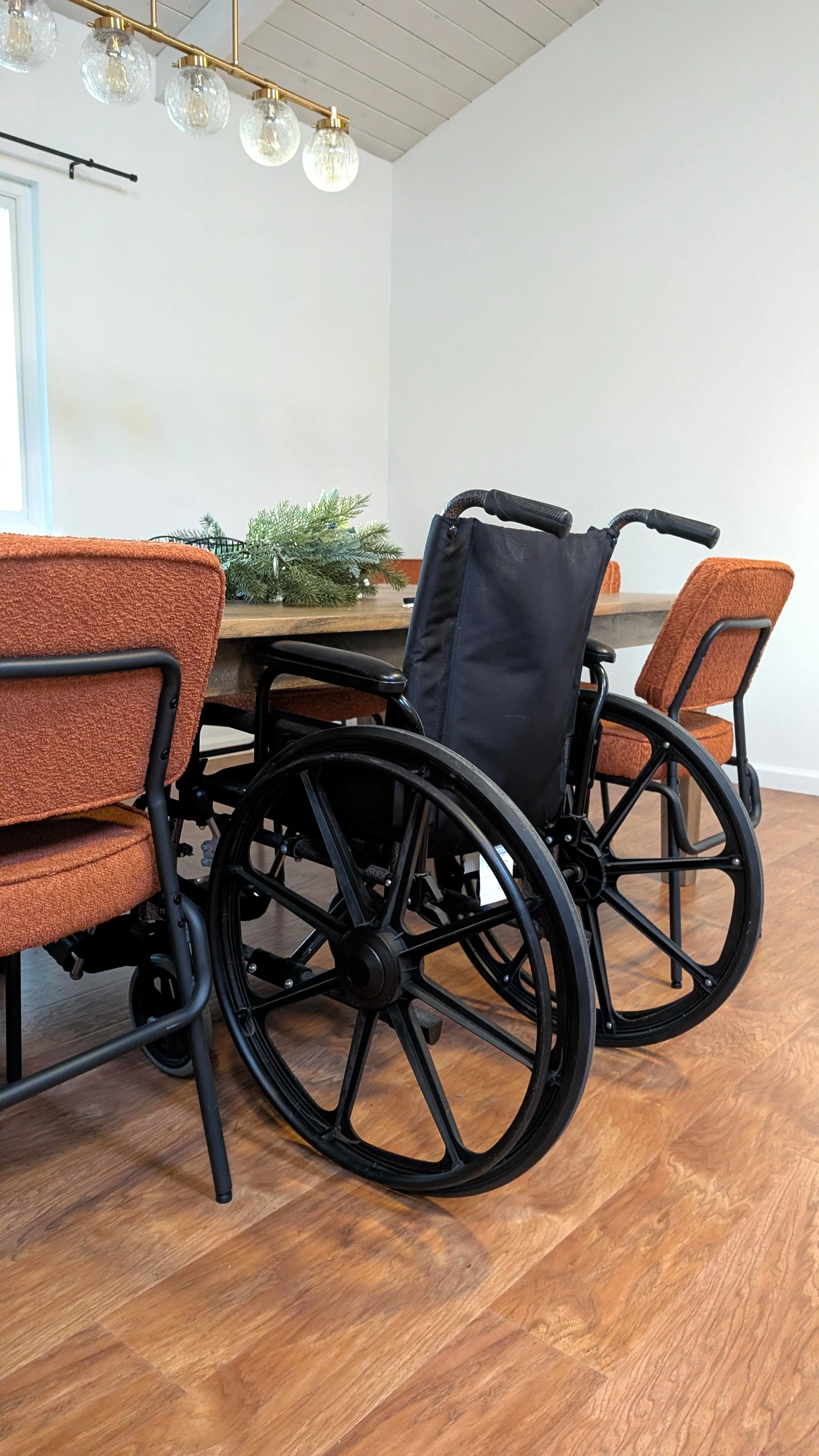 Interior room with a wooden dining table, orange upholstered chairs, a black wheelchair, and a decorative green plant on the table. White walls and a ceiling with a wooden panel design. A window with partially drawn black curtain and a modern chandel