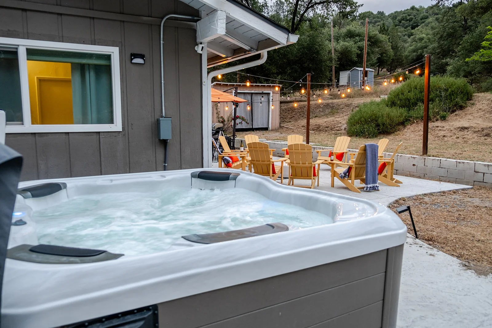 Outdoor patio with yellow Adirondack chairs, string lights, and a hot tub, set against a hillside with trees at dusk.