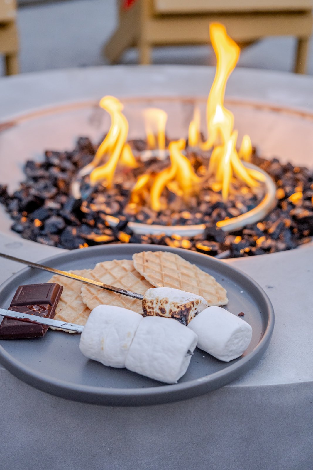 A plate of marshmallows, graham crackers, and chocolate near a fire pit with flames and black stones.