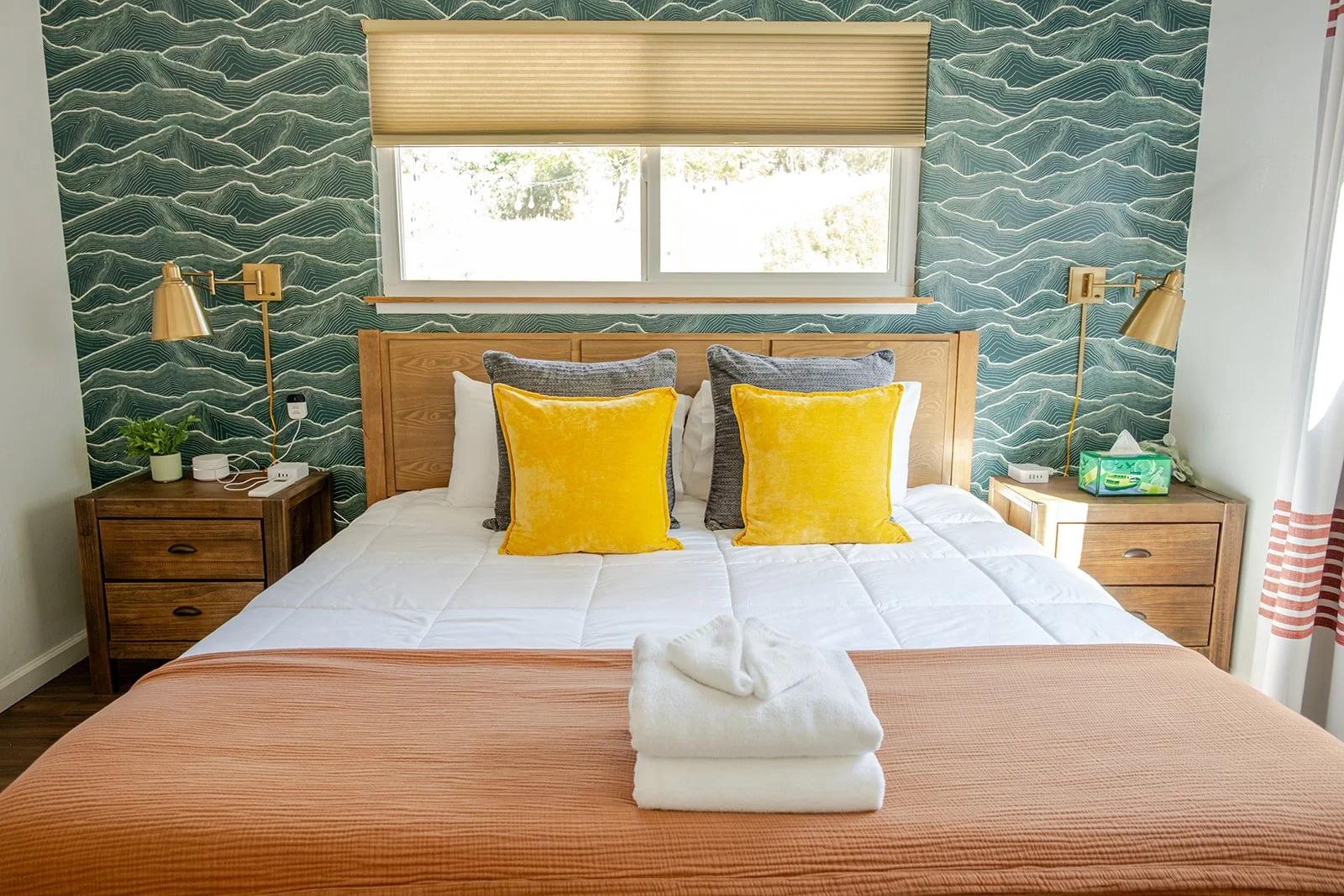 A neatly made bed with yellow and gray pillows, set in a bedroom with a patterned blue-green accent wall, wooden headboard, and matching nightstands with lamps, a plant, and a tissue box.