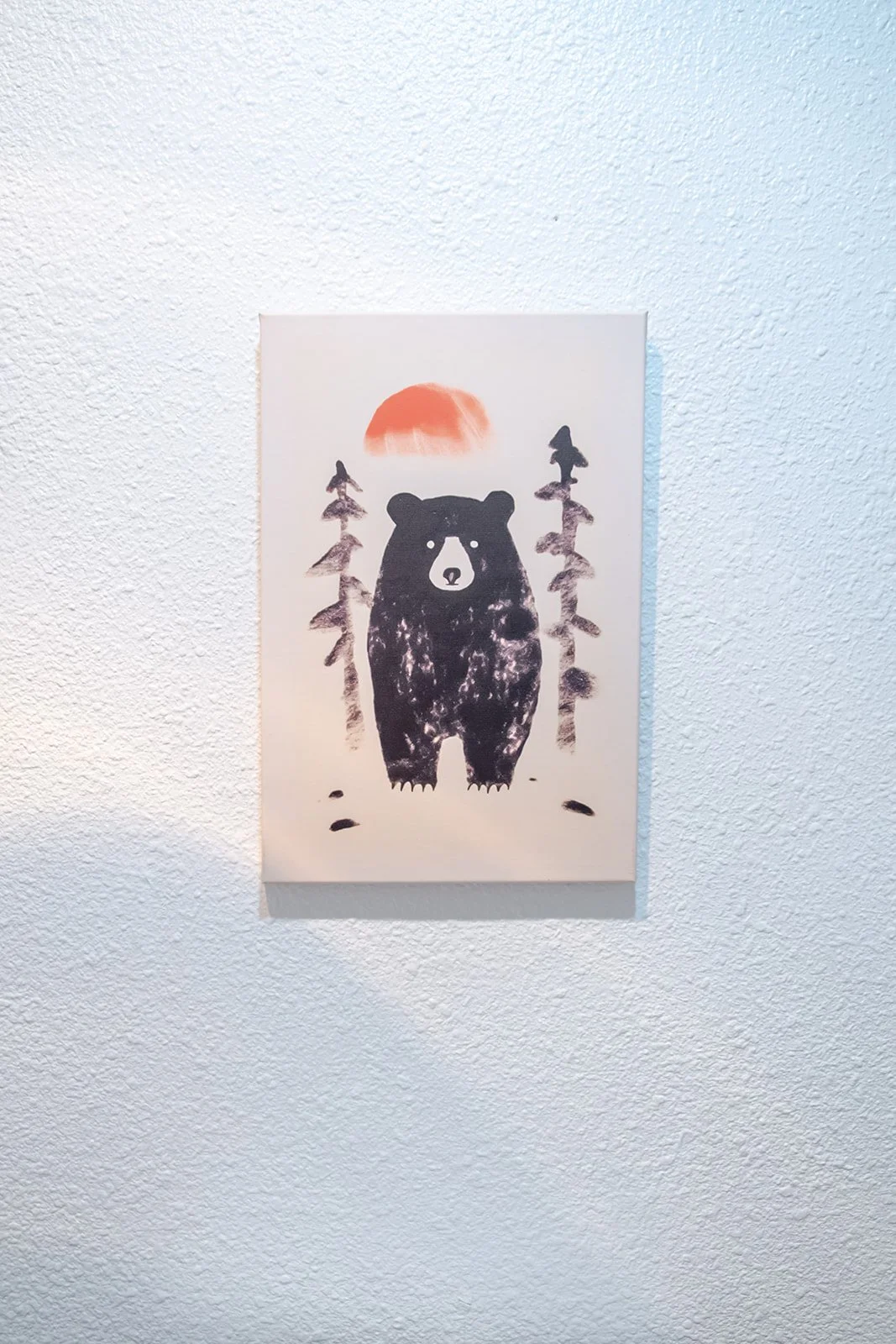 Acrylic painting of a bear standing amidst trees with a sun setting or rising in the background on a white textured wall.