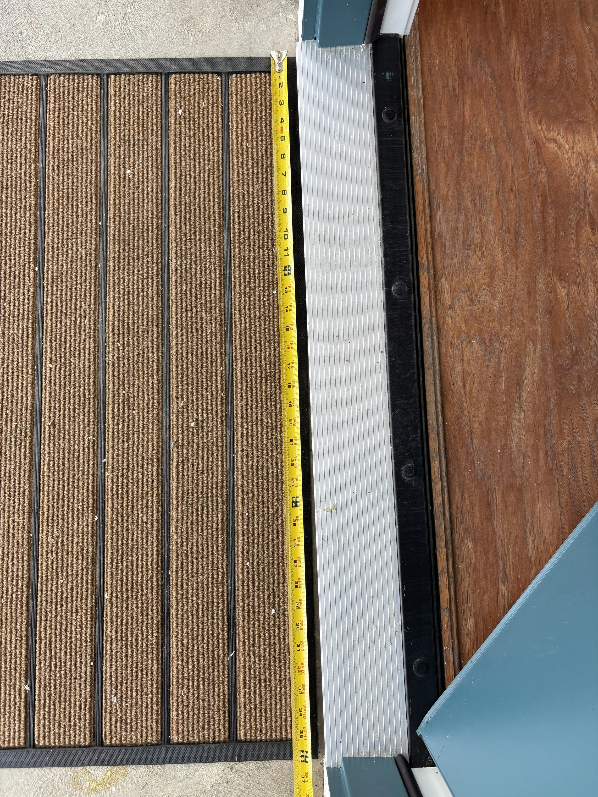 Close-up of a brown entry mat with black rubber border, a yellow measuring tape, and a metal door threshold on a wooden floor.