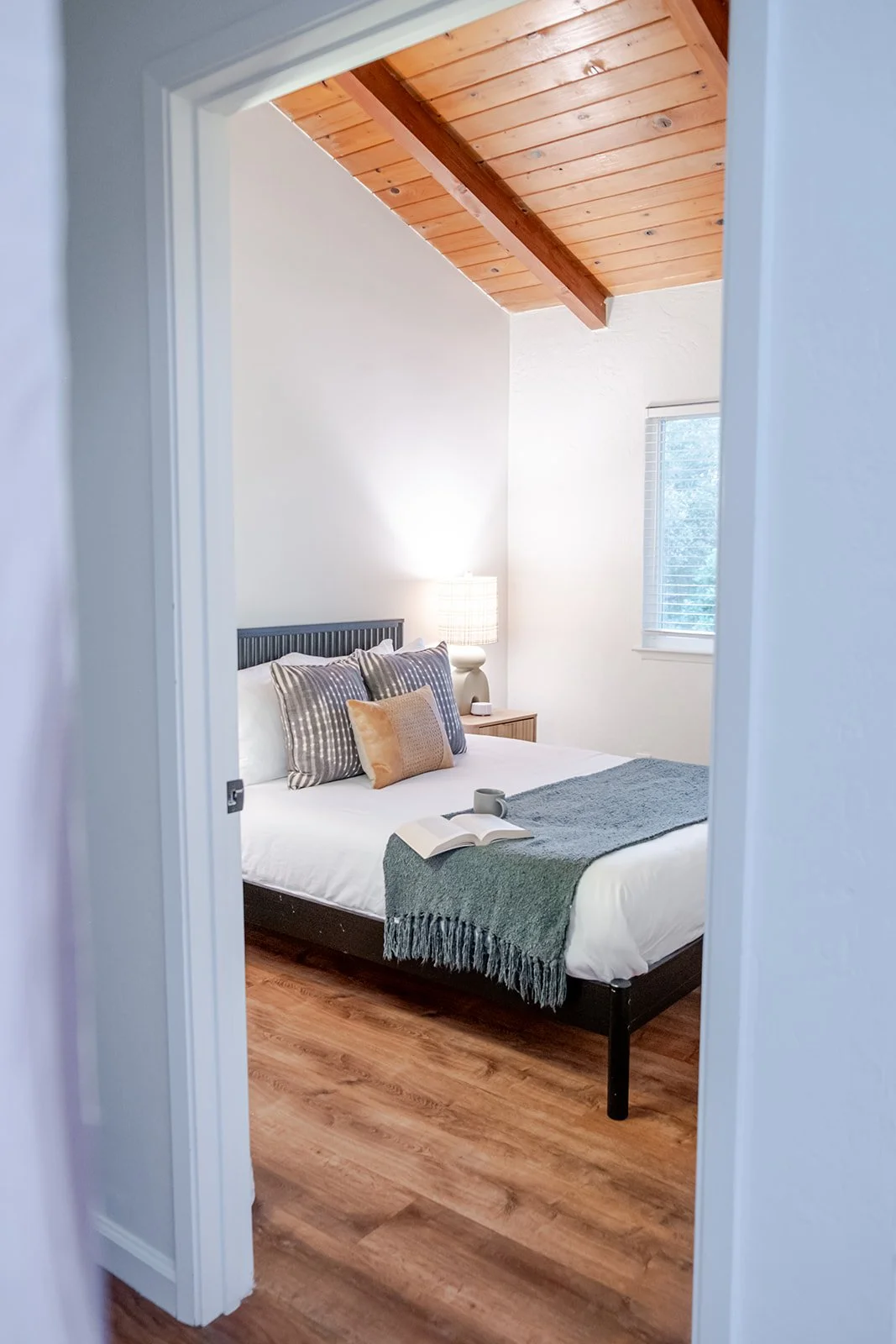 A cozy bedroom with a wooden ceiling, a bed with striped and solid pillows, a green throw blanket, a nightstand with a lamp, a window with blinds, and an open book with a cup on the bed.