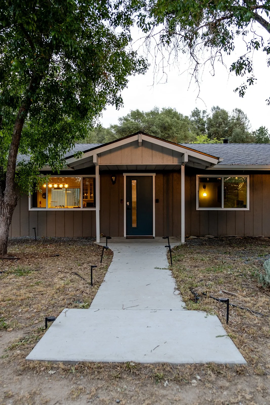 Front view of a single-story house with a concrete walkway, brown exterior, and large windows, surrounded by trees.