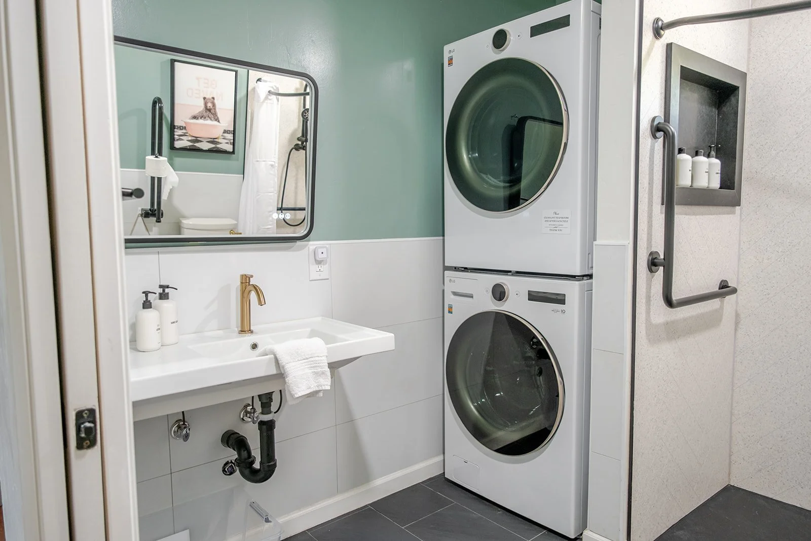 A  bathroom with a stacked washer and dryer, a small roll-under sink with a gold faucet, a large tilted smart mirror, and lotion and soap dispensers.