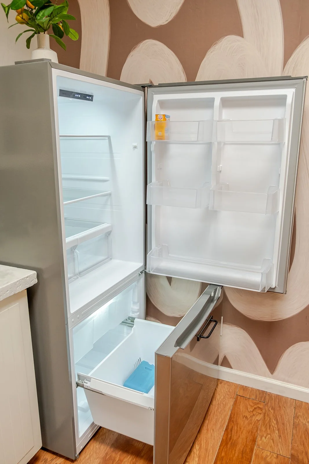 Open empty stainless steel refrigerator with a few shelves and transparent door compartments, situated in a room with wood flooring and patterned wall.