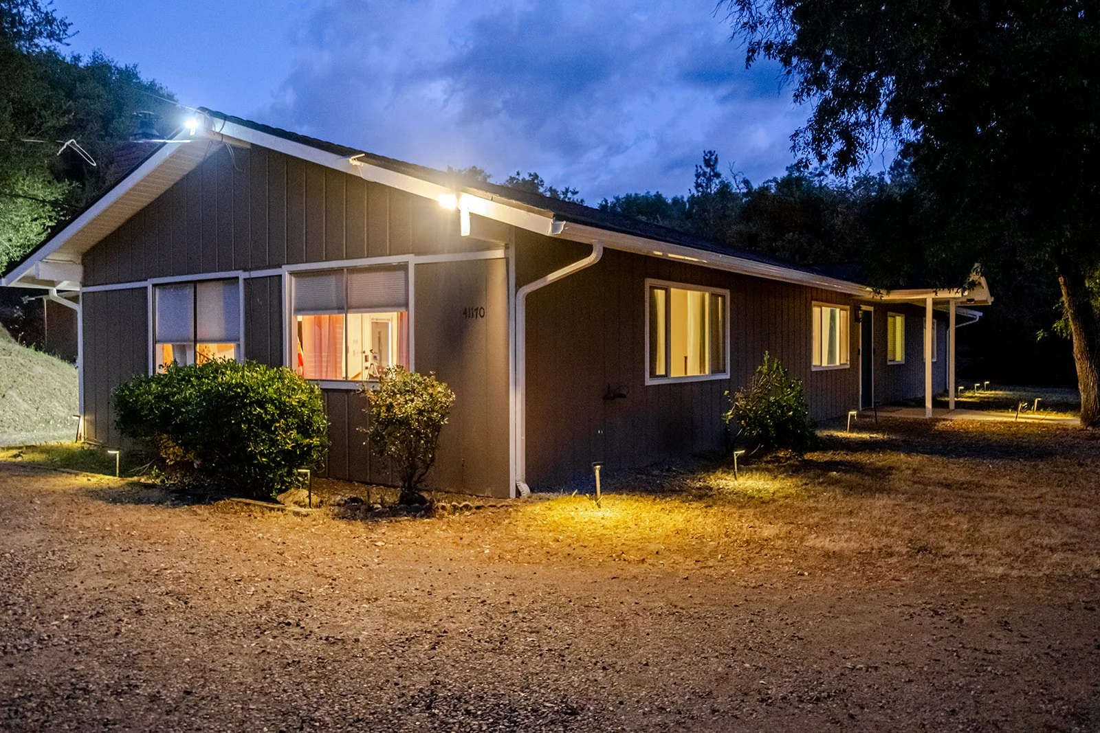 A house at dusk with lights on inside, dark green exterior, bushes in front, and a gravel driveway, situated in a natural landscape with trees and a cloudy sky.