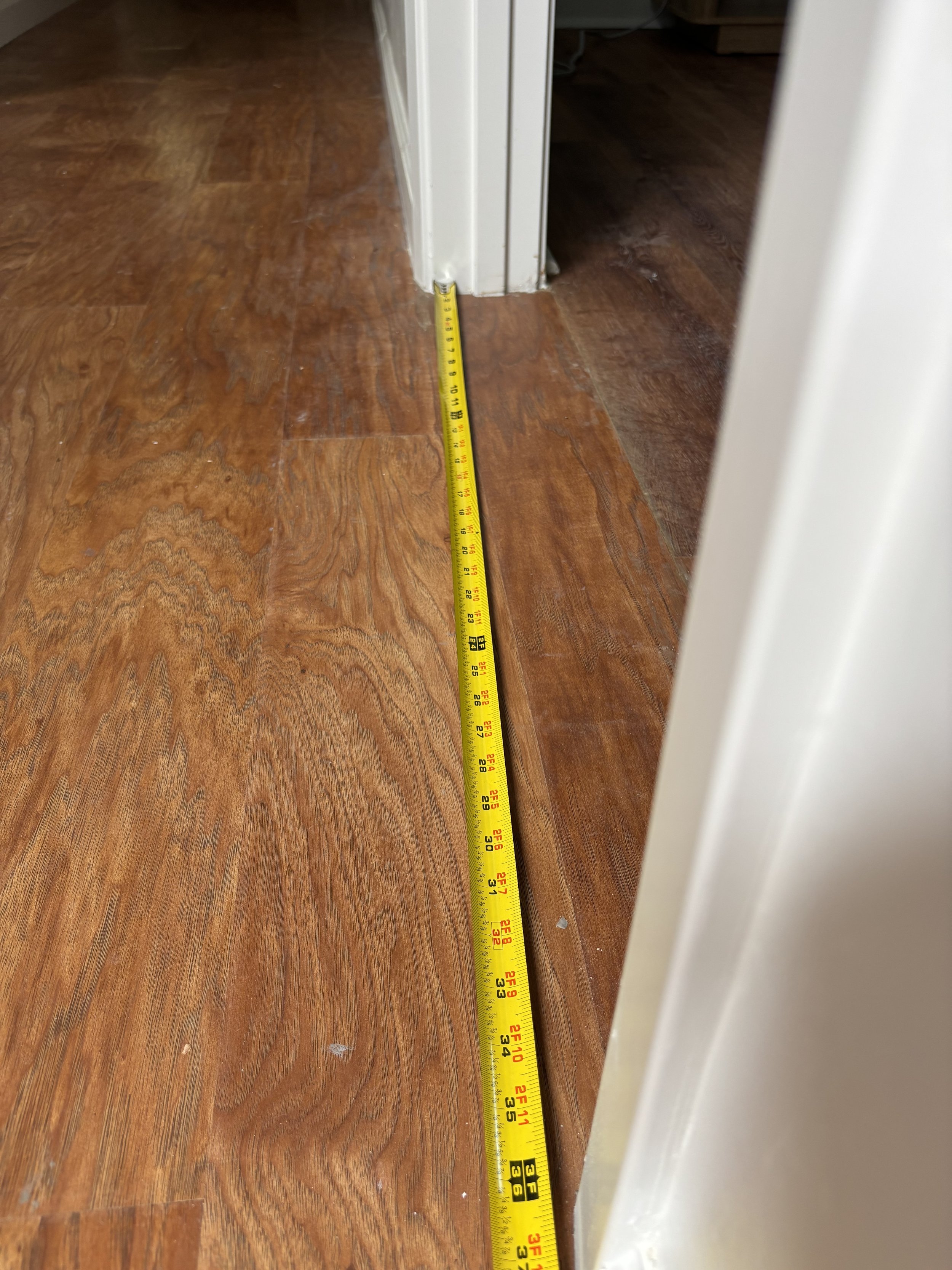 A yellow measuring tape stretched across wooden floor, measuring the distance from the doorway to the wall.
