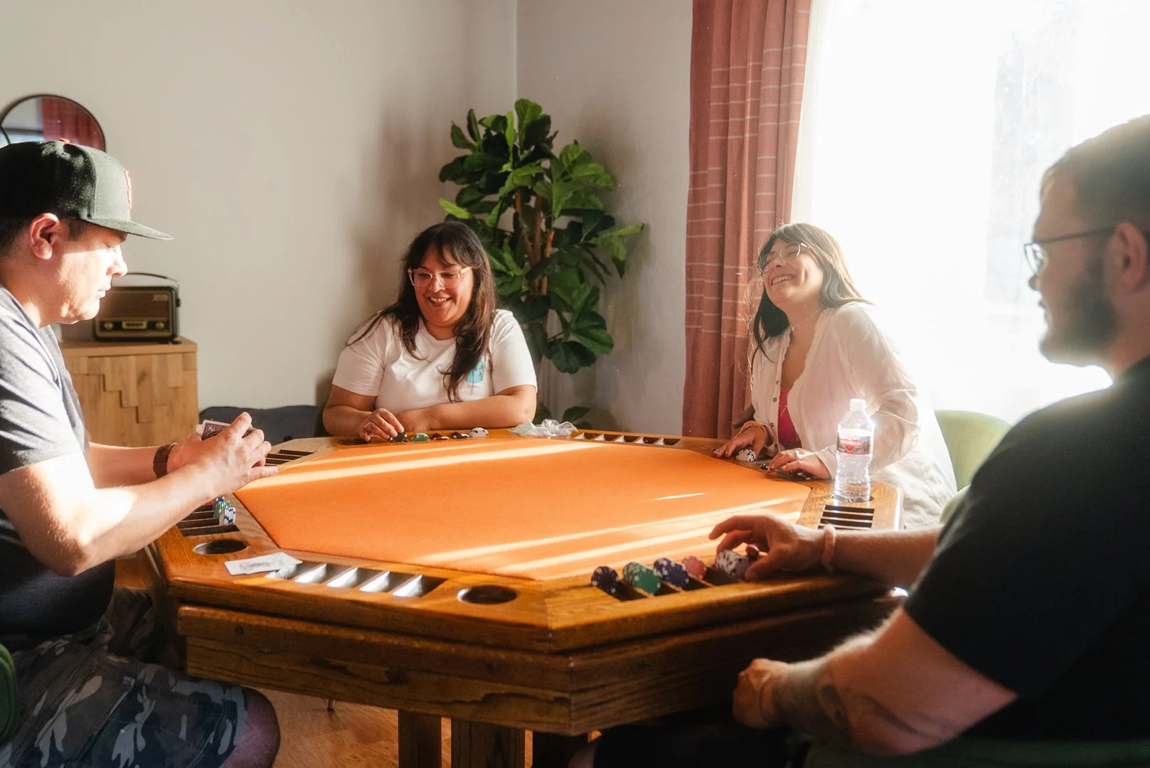Four people sitting around a wooden table playing a game in a sunlit room, smiling and enjoying each other's company.