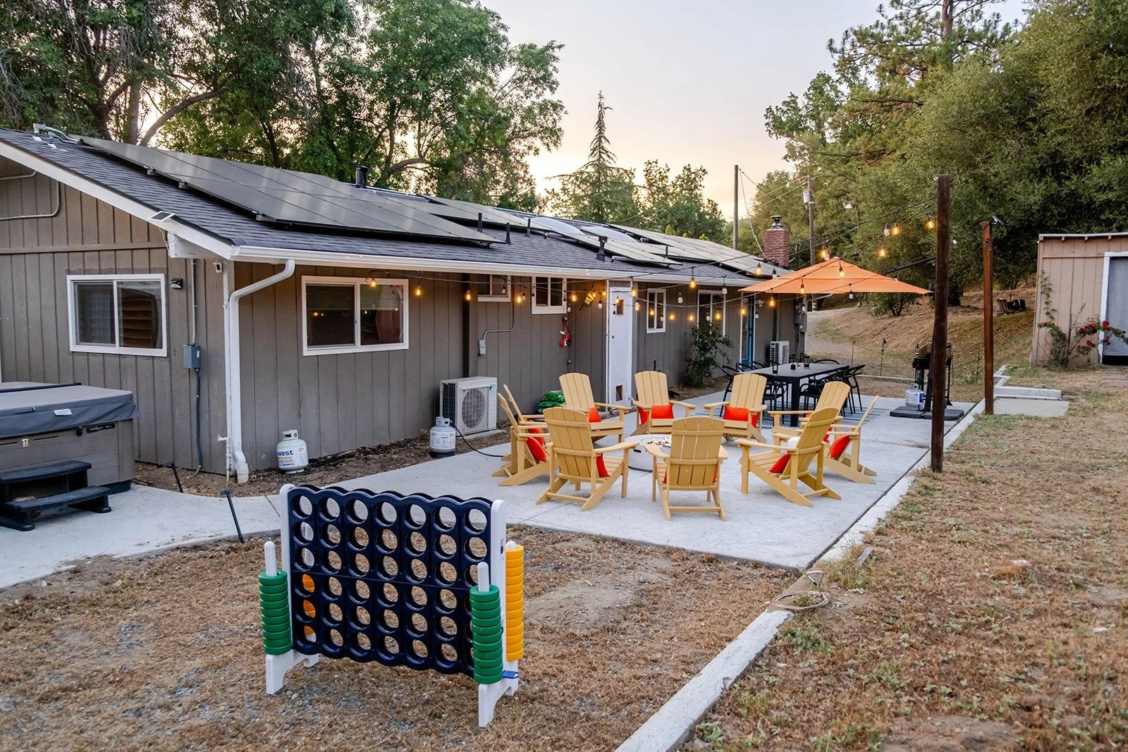Outdoor backyard patio area with a concrete surface, featuring yellow Adirondack chairs with red cushions arranged around a fire pit, a black dining table with chairs, string lights overhead, a large orange patio umbrella, a small game called Connect