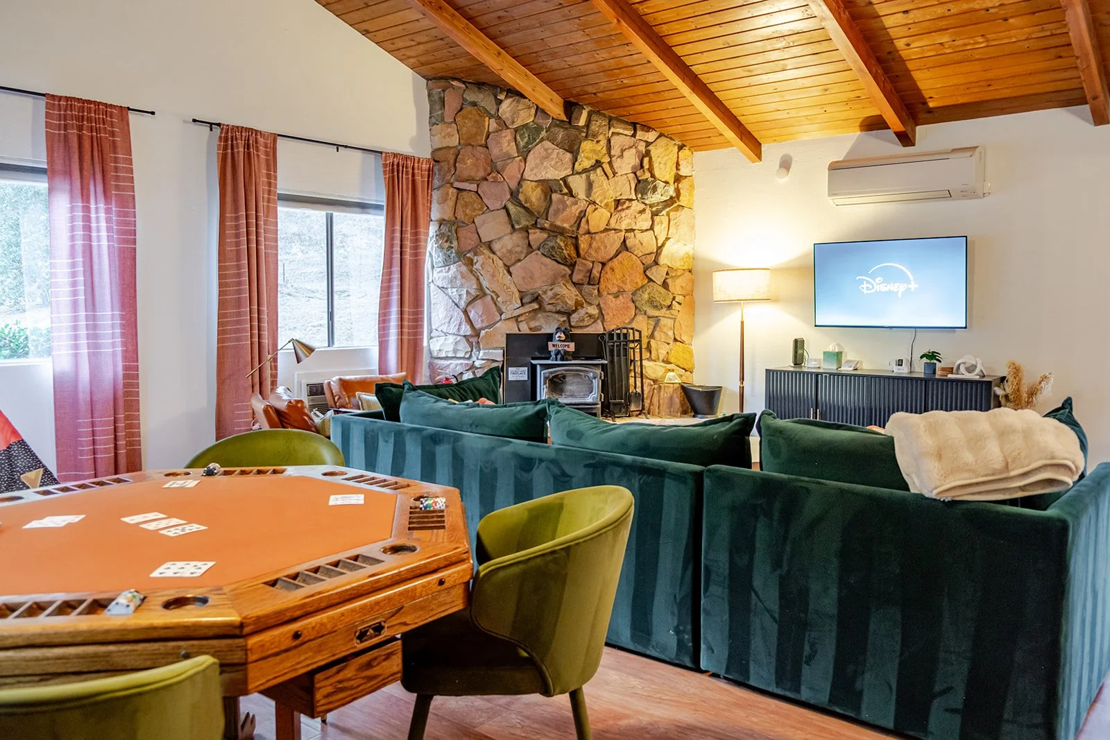 Living room with a wood-paneled ceiling, a stone fireplace, a flat-screen TV, a beige armchair with a blanket, a green sofa, and a round poker game table with four green chairs.