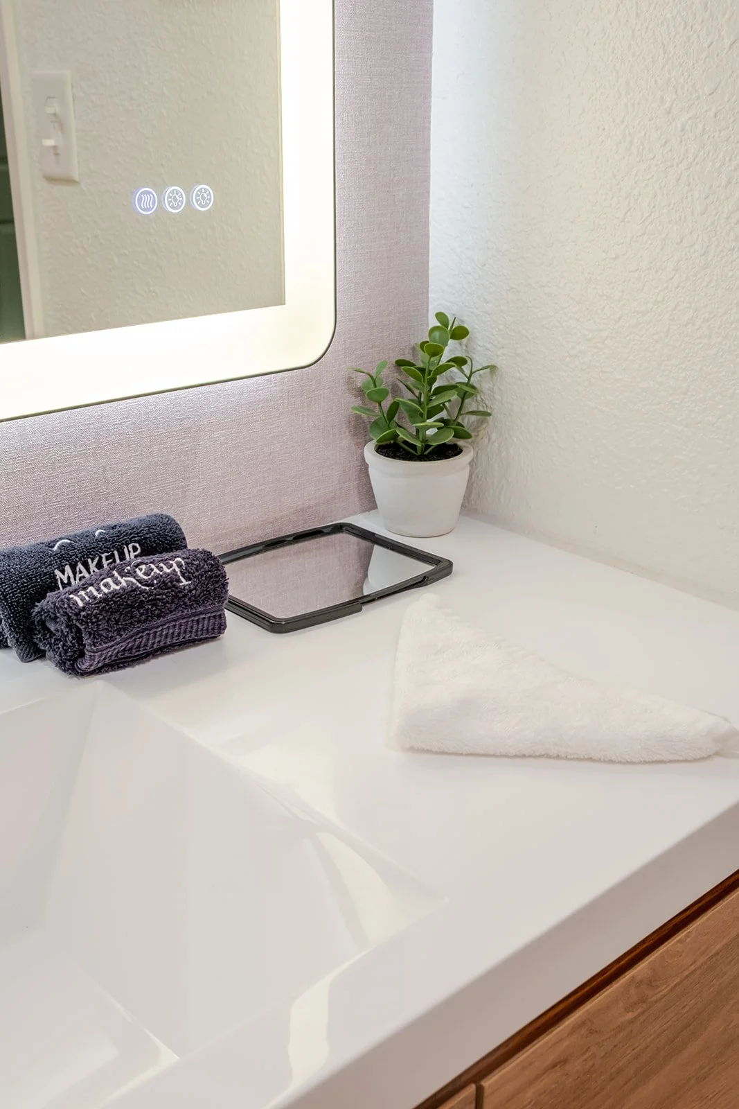 Bathroom countertop with a mirror, a small potted plant, a stack of dark makeup towels, a small mirror, a folded white towel, and a pink wall behind.