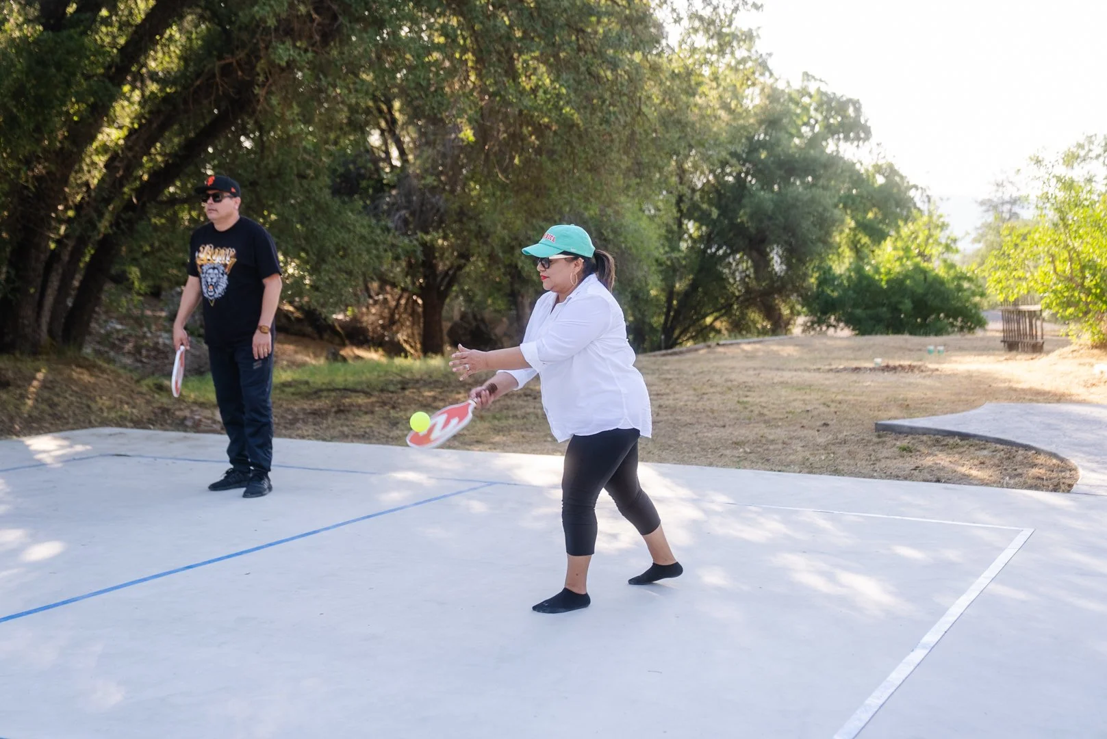 Two people playing pickleball outdoors on a court surrounded by trees, with one woman preparing to hit a yellow ball with her paddle.