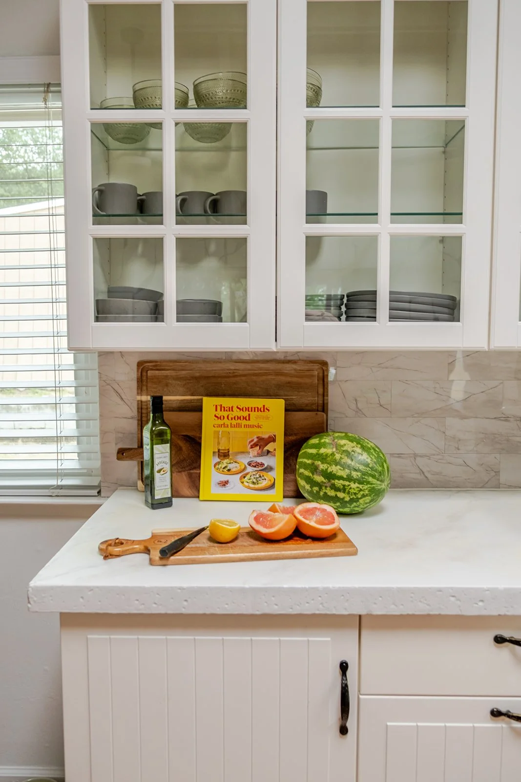 Kitchen countertop with a watermelon, a lemon, grapefruit halves, a bottle of olive oil, a cutting board, a knife, a cookbook titled 'That Sounds So Good,' and a wooden tray with glass bowls stored in the glass-front cabinet above.