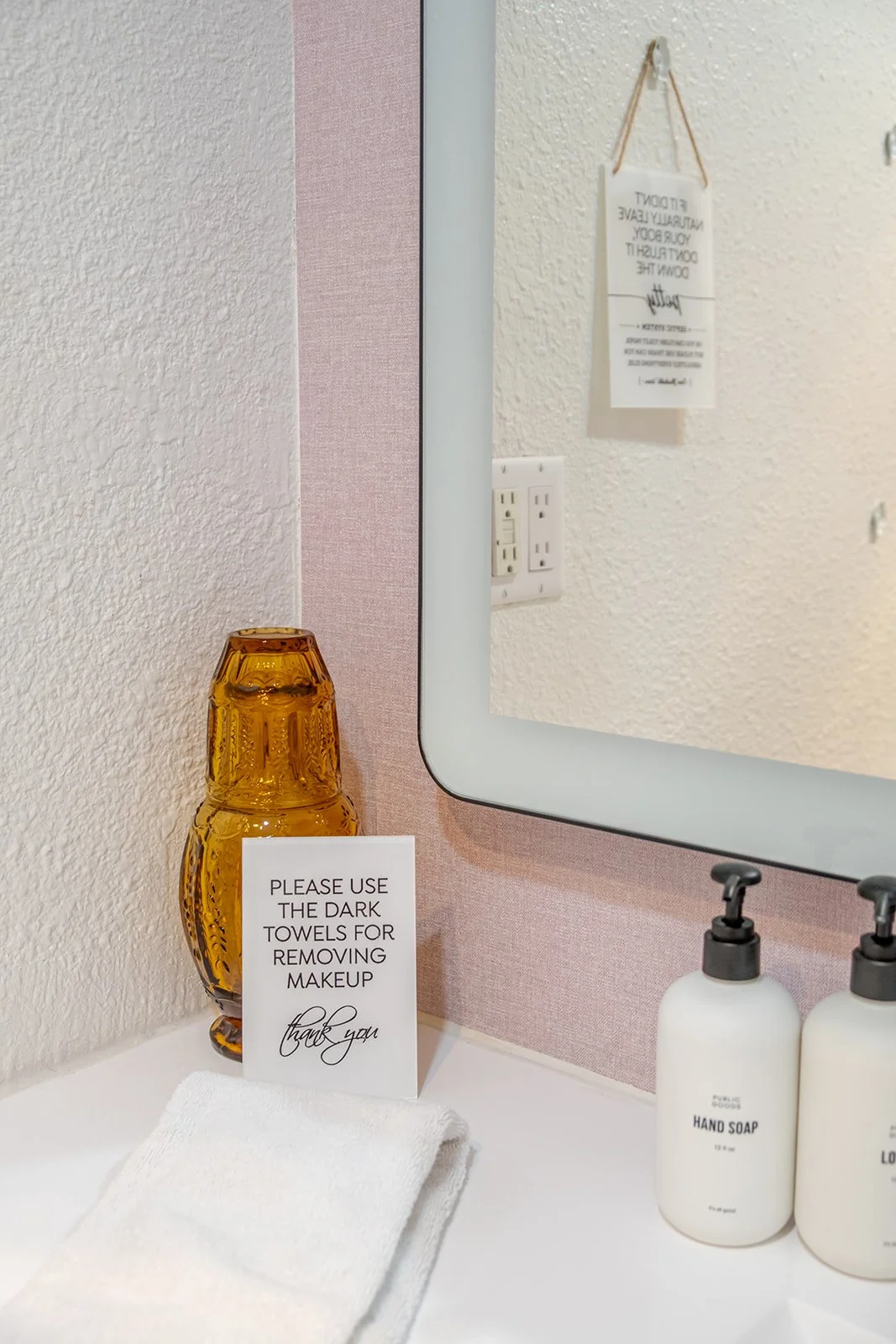 Bathroom sink area with a mirror, amber glass vase, white towel, sign asking to use dark towels for makeup removal, white hand soap bottle, and background wall with electrical outlets and a decorative sign.